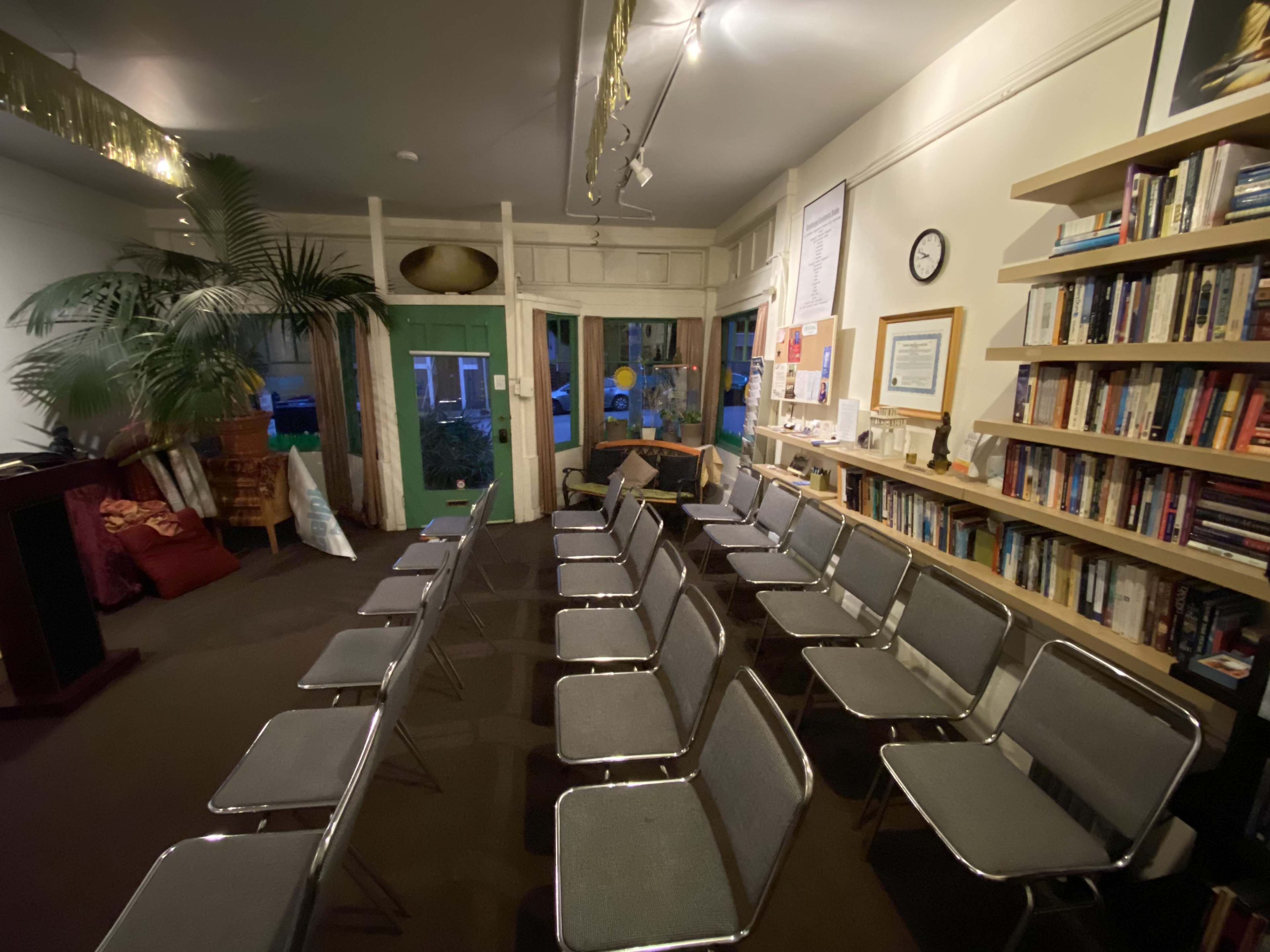 A room with several metal-framed chairs arranged in rows facing a lectern, alongside a bookshelf and large windows with green doors.