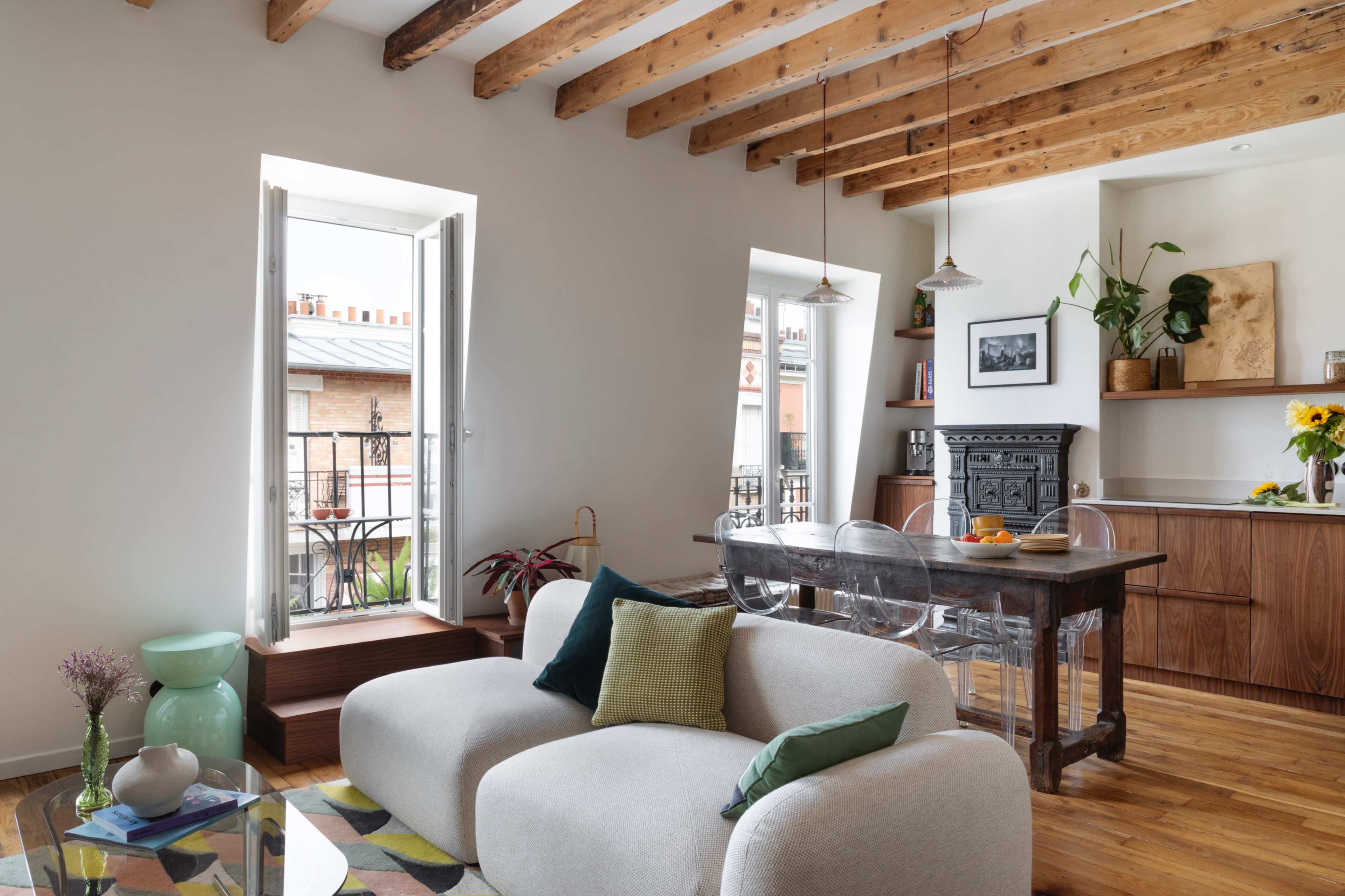 The image shows a modern living space with exposed wooden beams, a cozy sofa, a dining area with a wooden table, and large windows that open to a balcony.