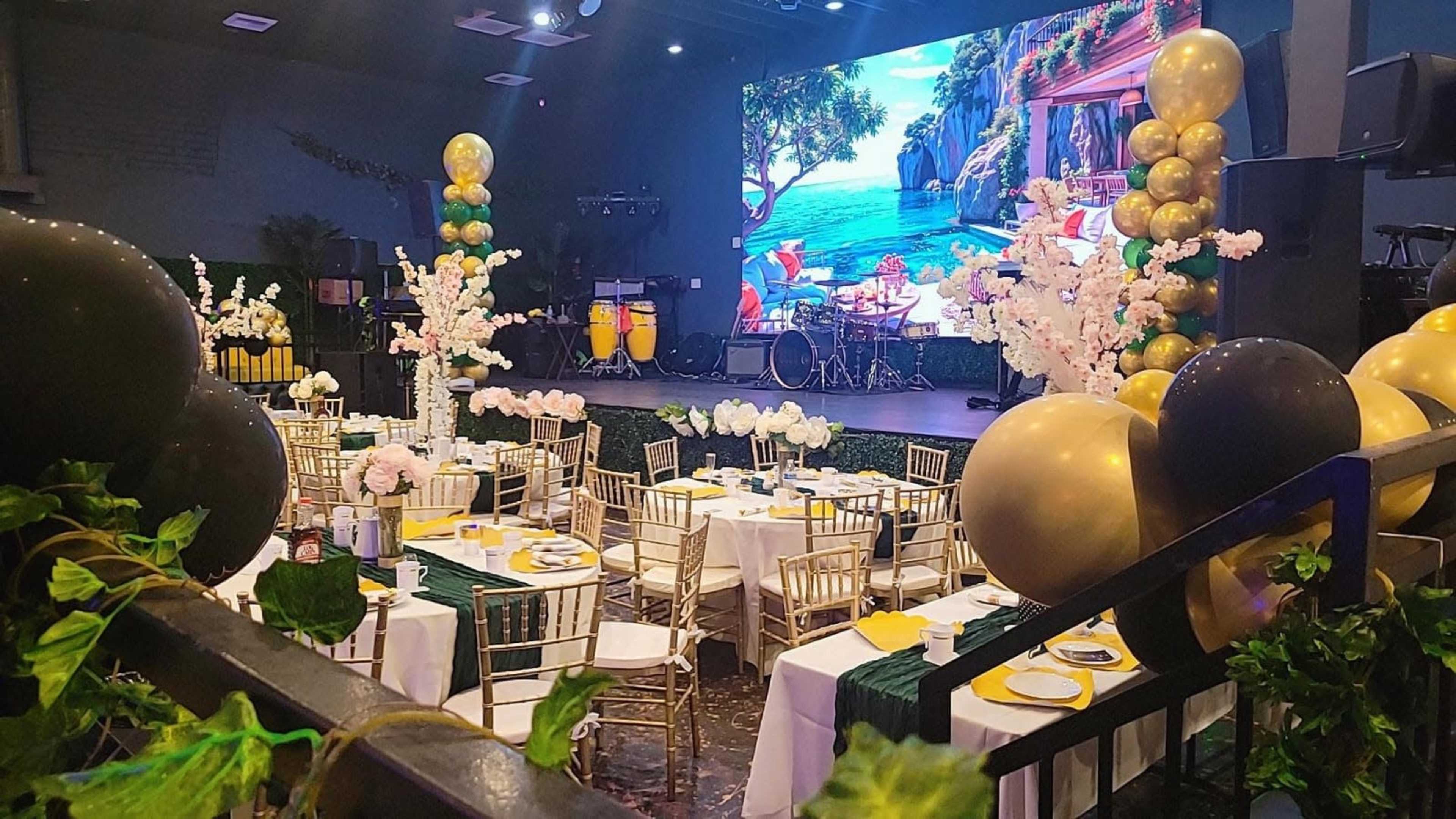 The image shows a decorated event space with round tables set for a celebration, featuring floral centerpieces and golden and black balloon arrangements, while a large screen displays a vibrant outdoor scene in the background.