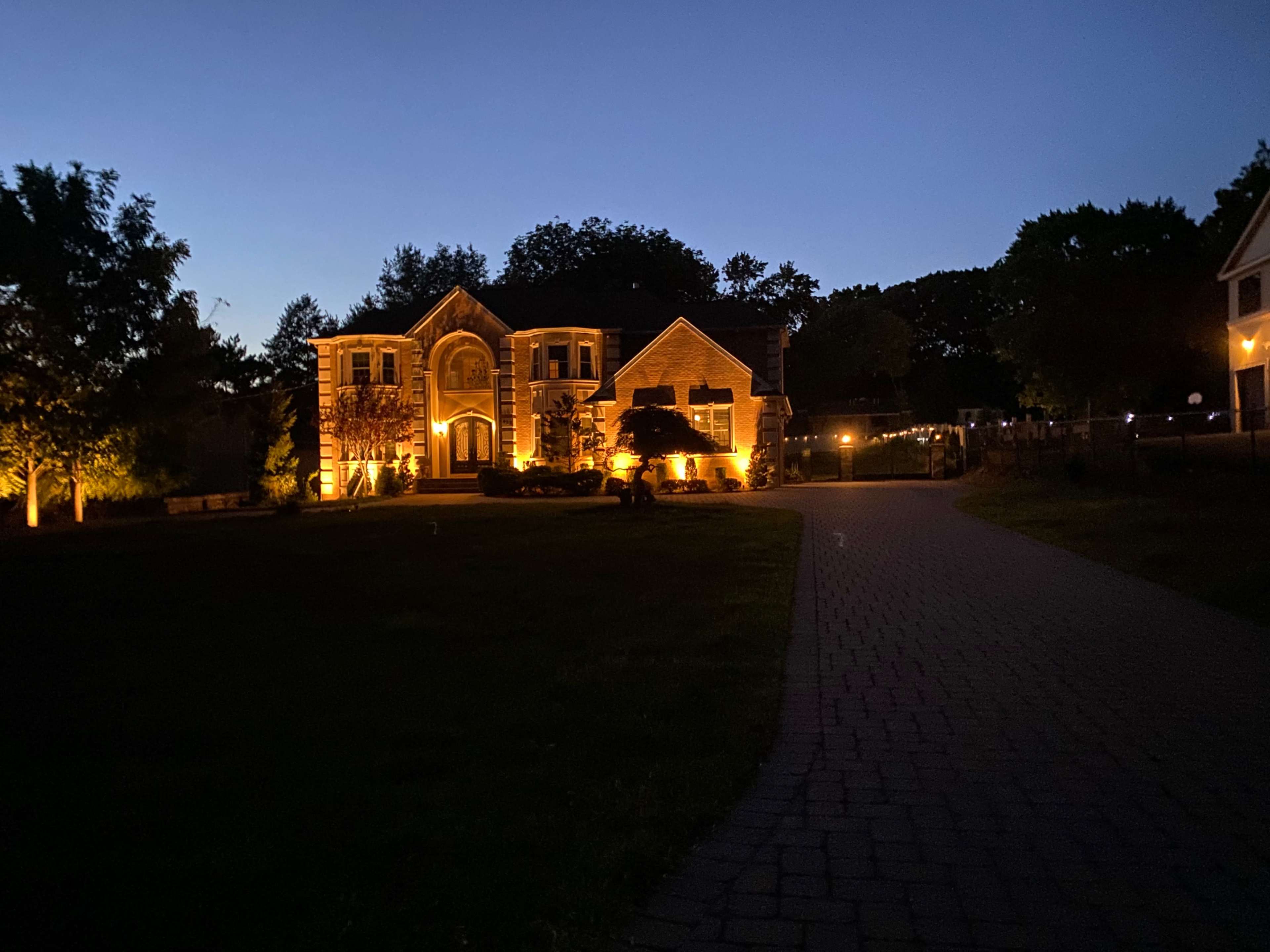 A well-lit two-story house is surrounded by trees and a pathway during twilight.