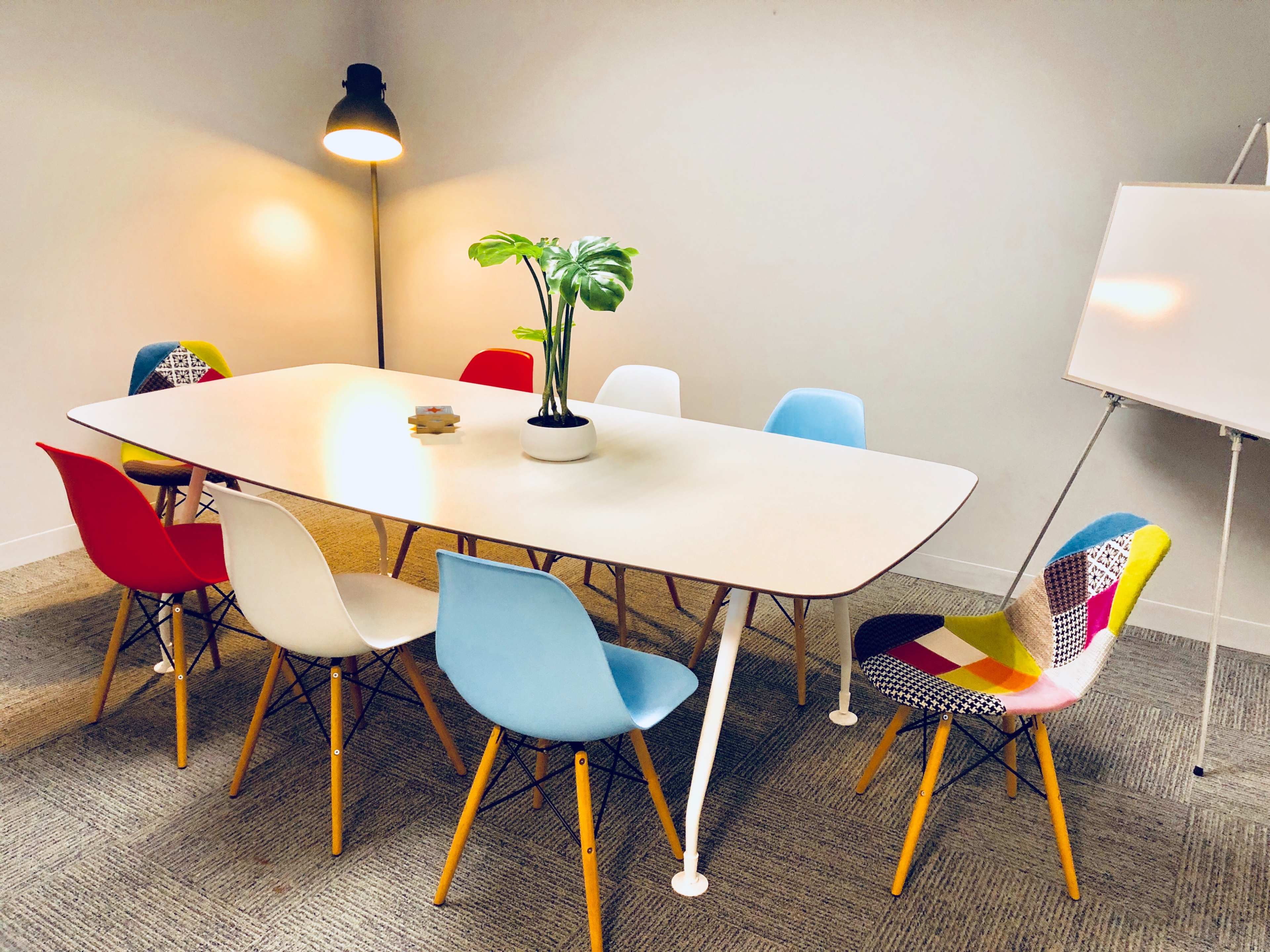 A modern conference room features a long table surrounded by eight colorful chairs and a floor lamp, with a potted plant in the center.