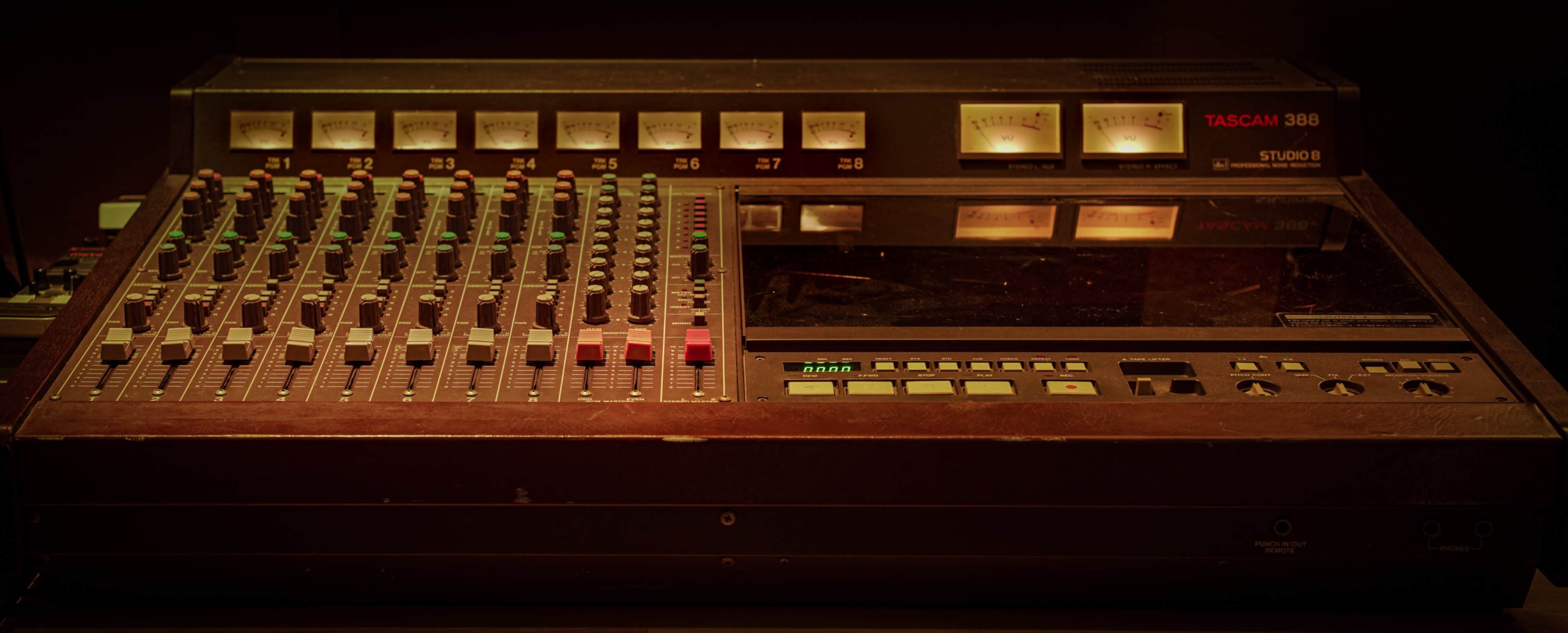 The image shows a vintage Tascam 388 audio mixer with multiple knobs, faders, and meters for audio level monitoring.