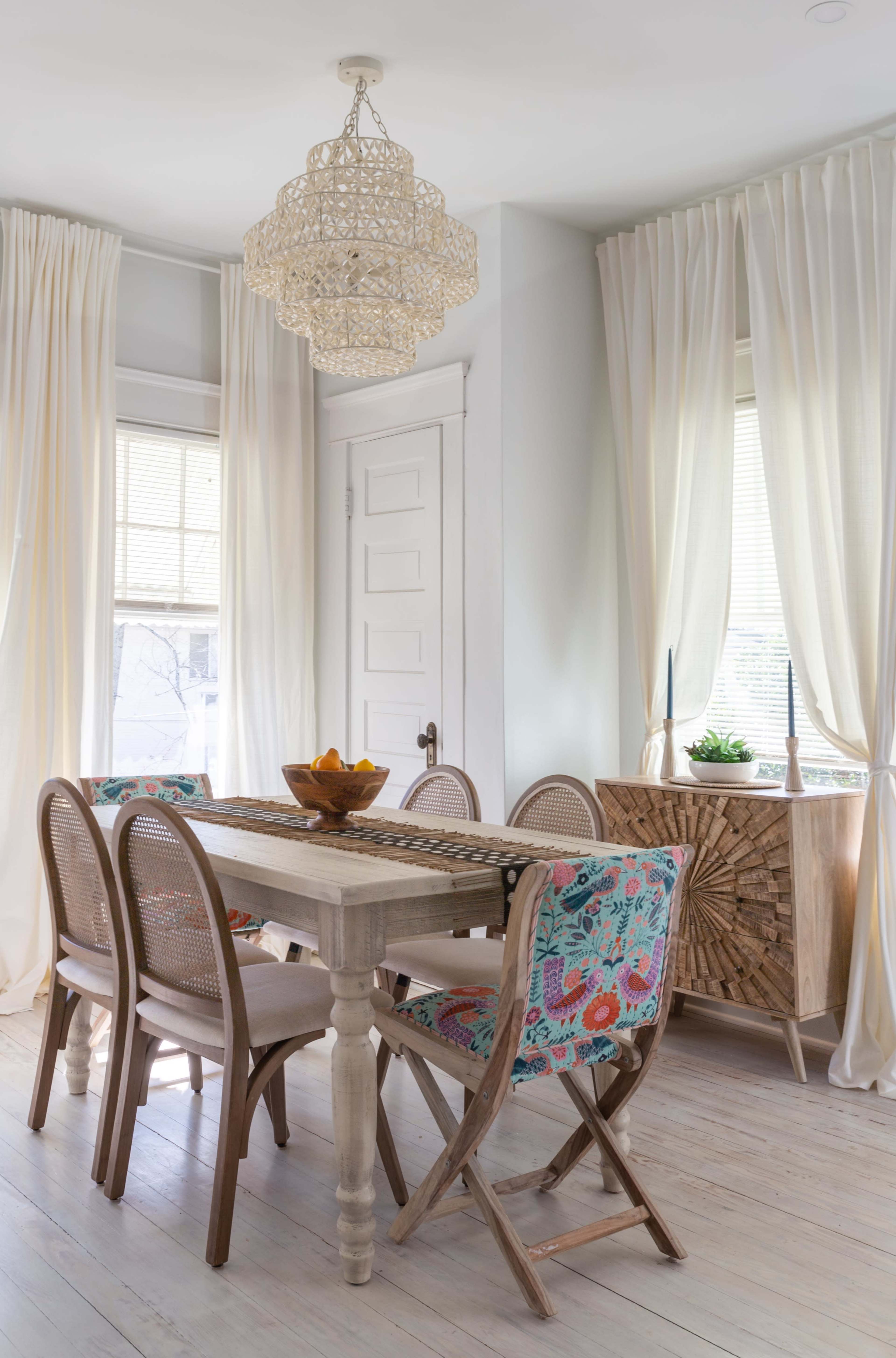 A dining room features a wooden table surrounded by four chairs with patterned cushions, a woven chandelier above, and large windows draped with white curtains.