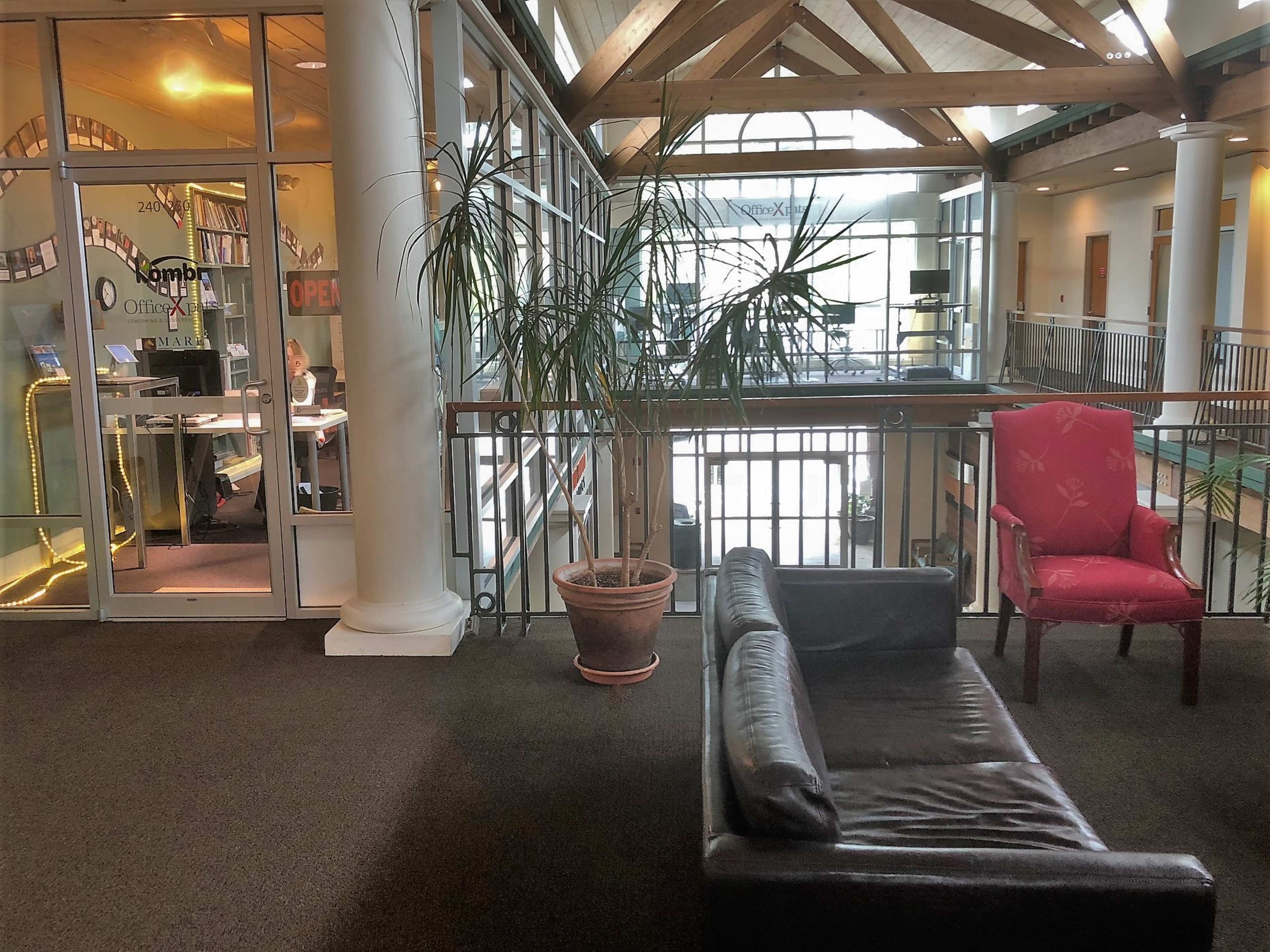 A modern office interior with large windows, a couch and a potted plant on a balcony overlooking a workspace.