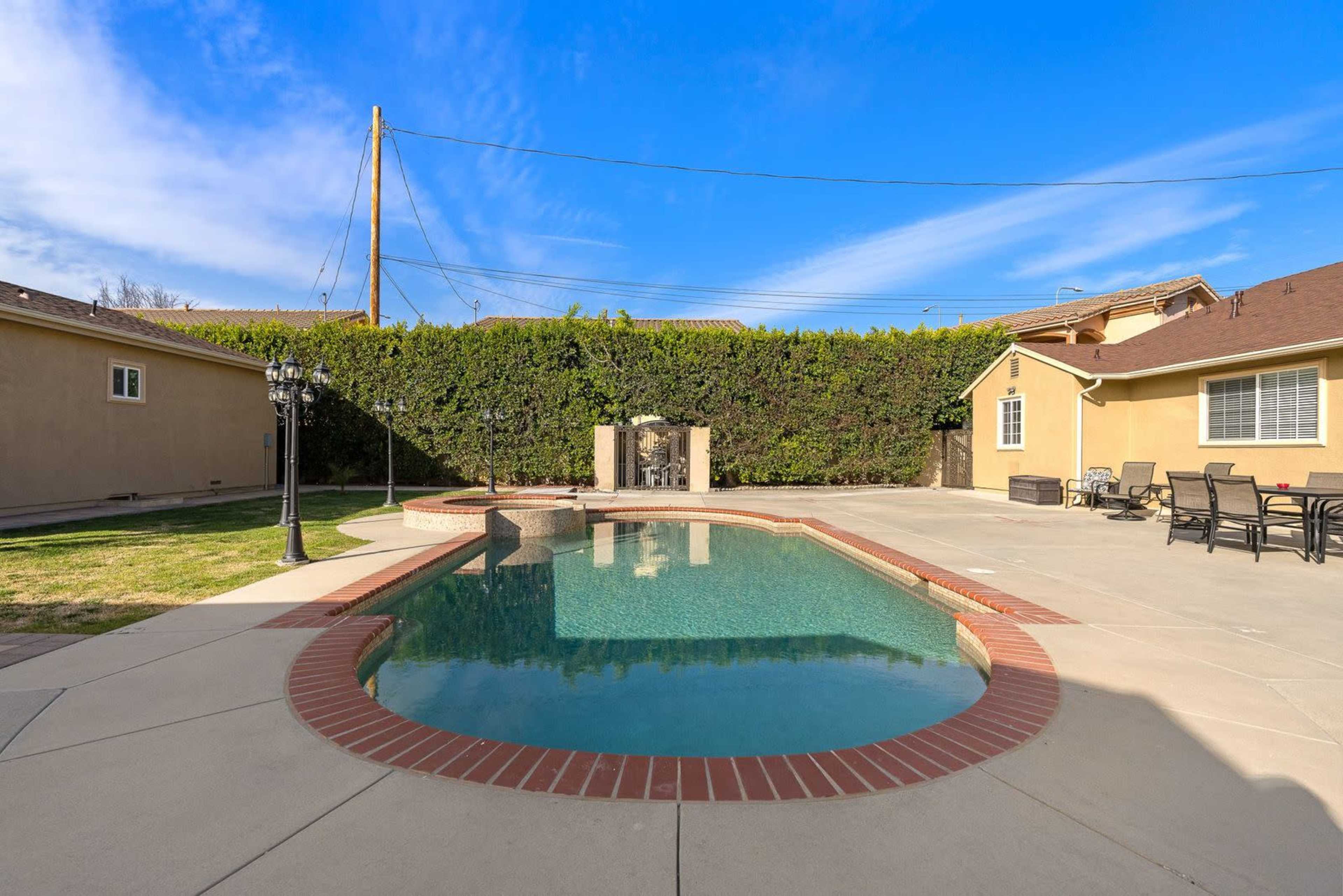 The image shows a backyard featuring a swimming pool with a brick border, surrounded by neatly trimmed hedges and two houses with patio furniture nearby.