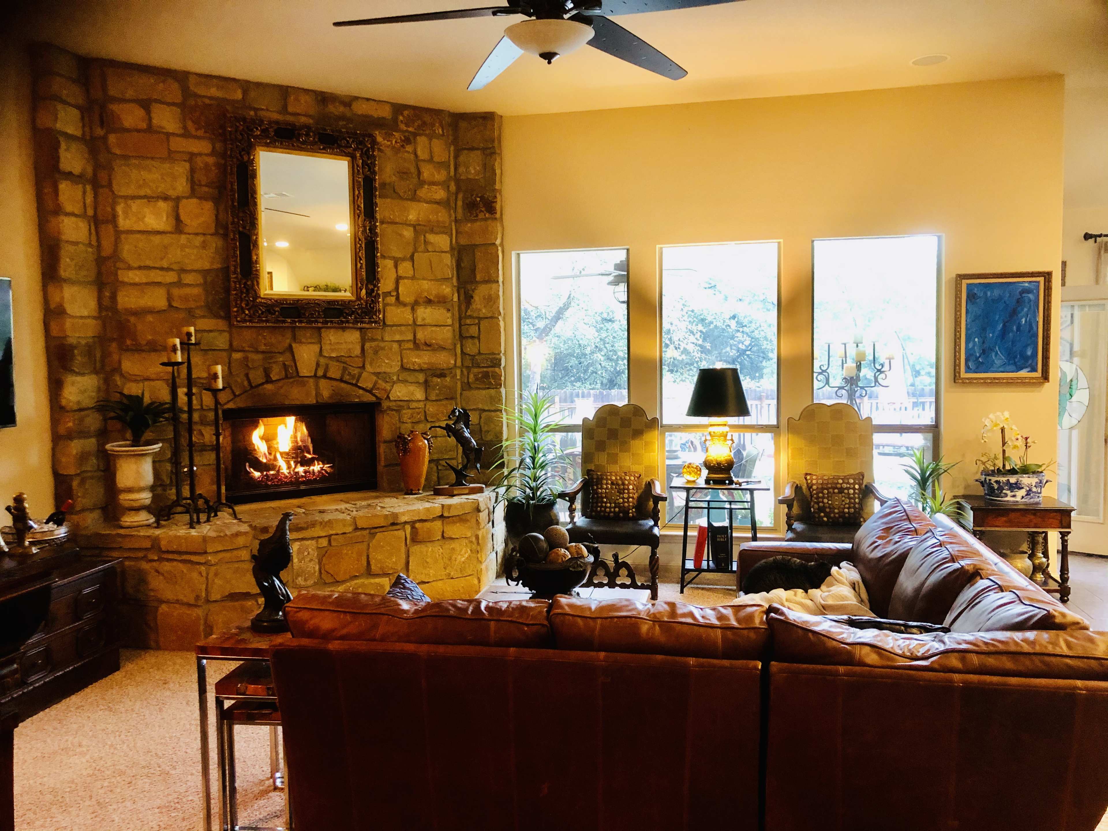 A cozy living room features a stone fireplace with a fire burning, surrounded by large windows, comfortable seating, and various decorative elements.