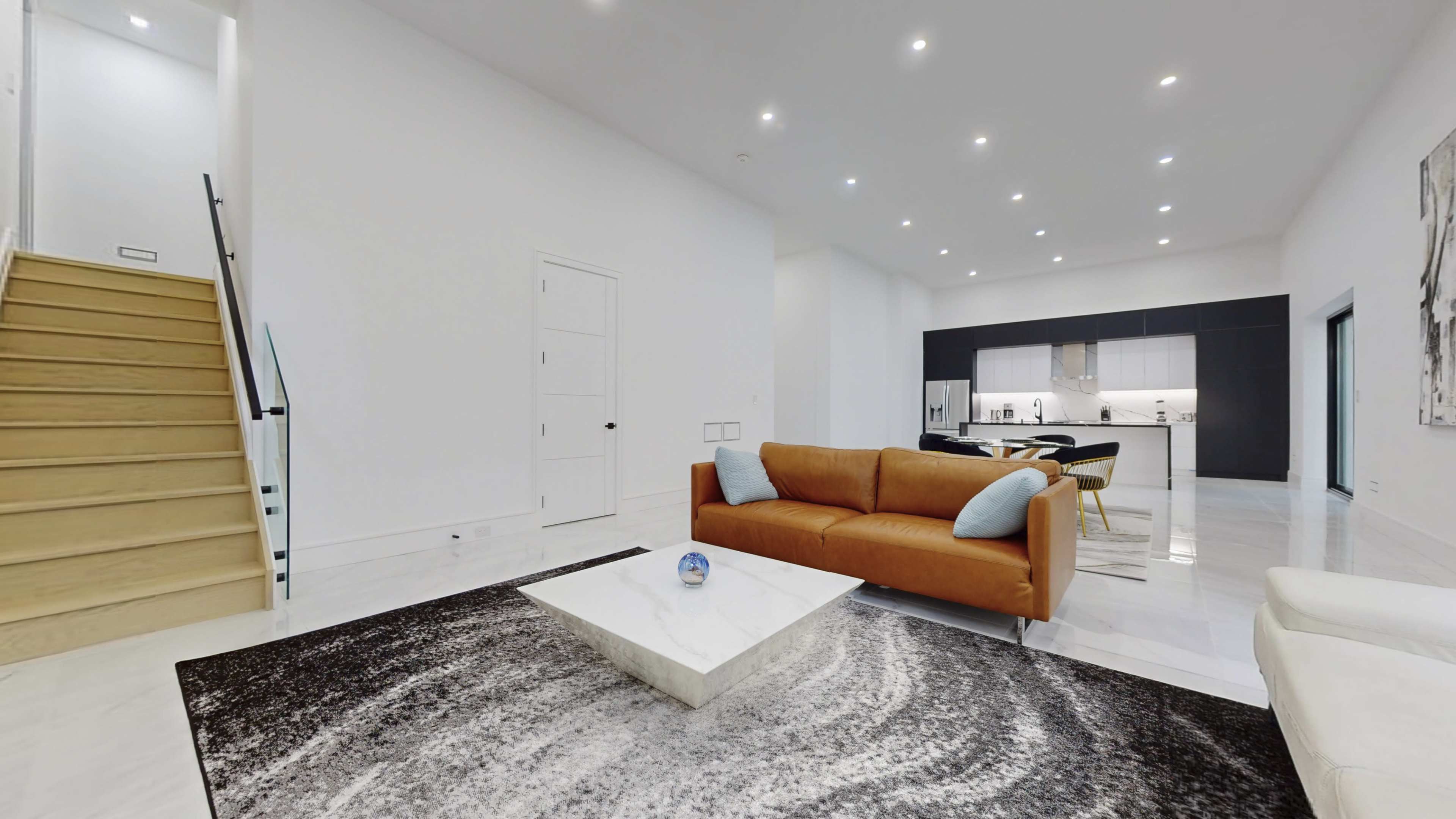 A modern living room with a brown sofa, a marble coffee table, and a staircase leading to an upper level, with a kitchen visible in the background.