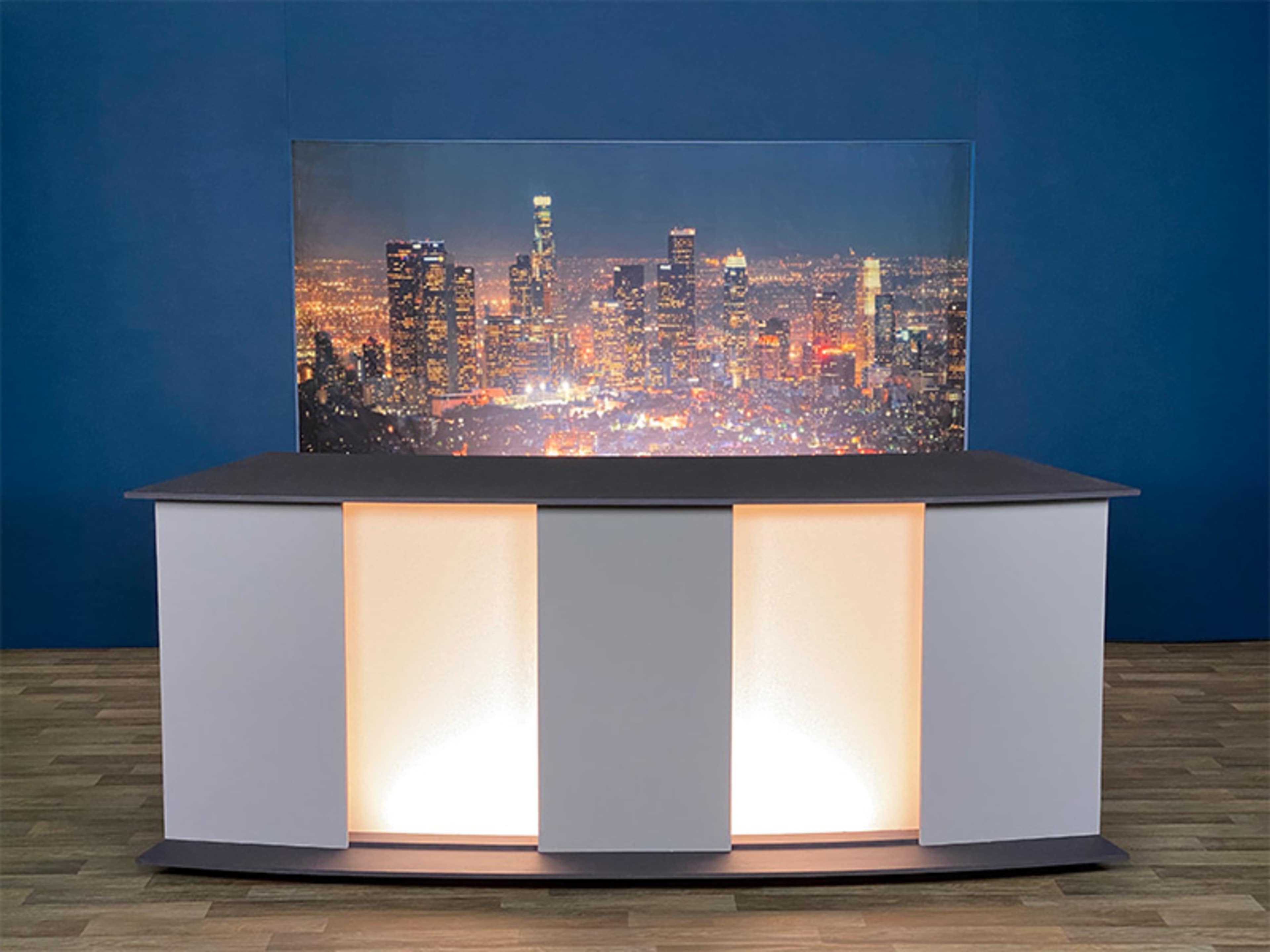 A modern presentation stage features a sleek counter with illuminated side panels and a backdrop of a city skyline at night.