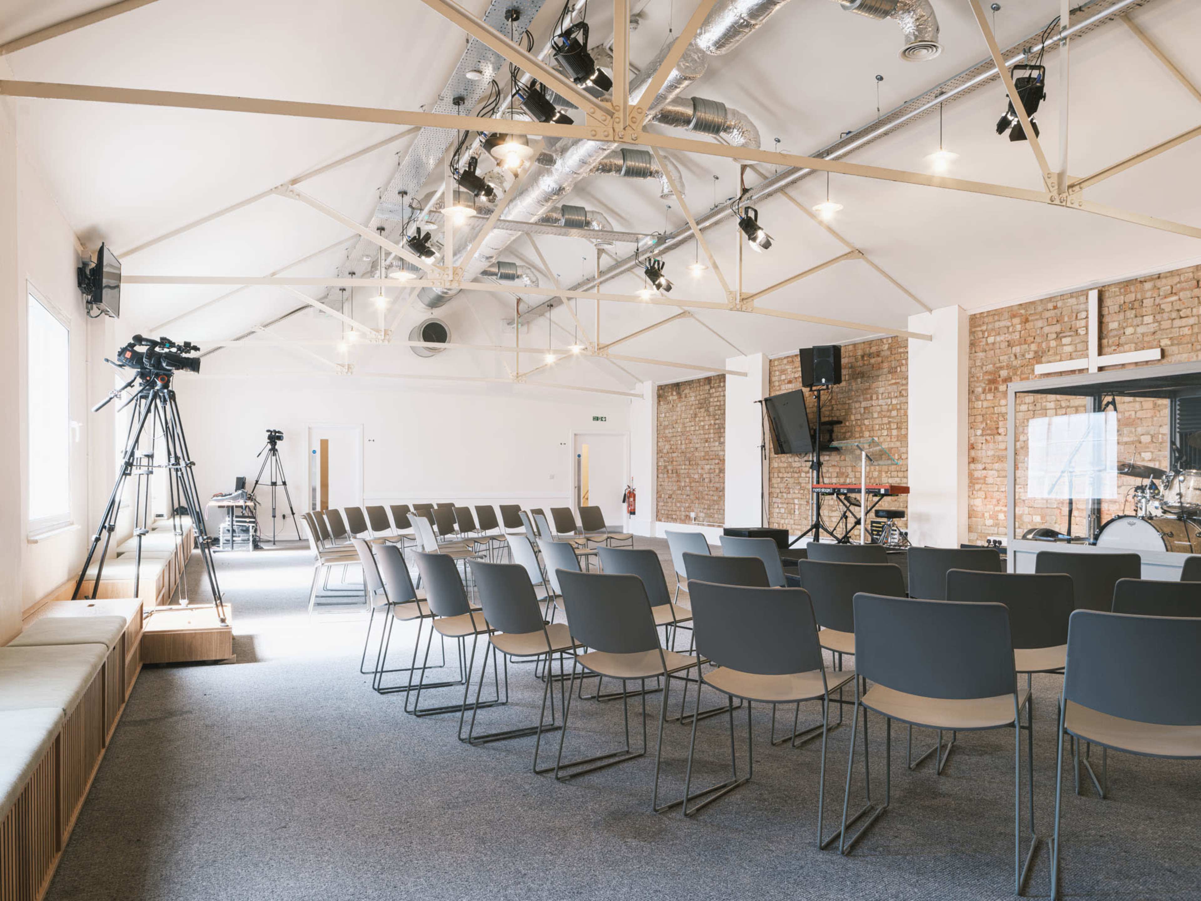 A spacious, modern interior of a seating area set up for an event, featuring rows of chairs, a stage, and audio-visual equipment.