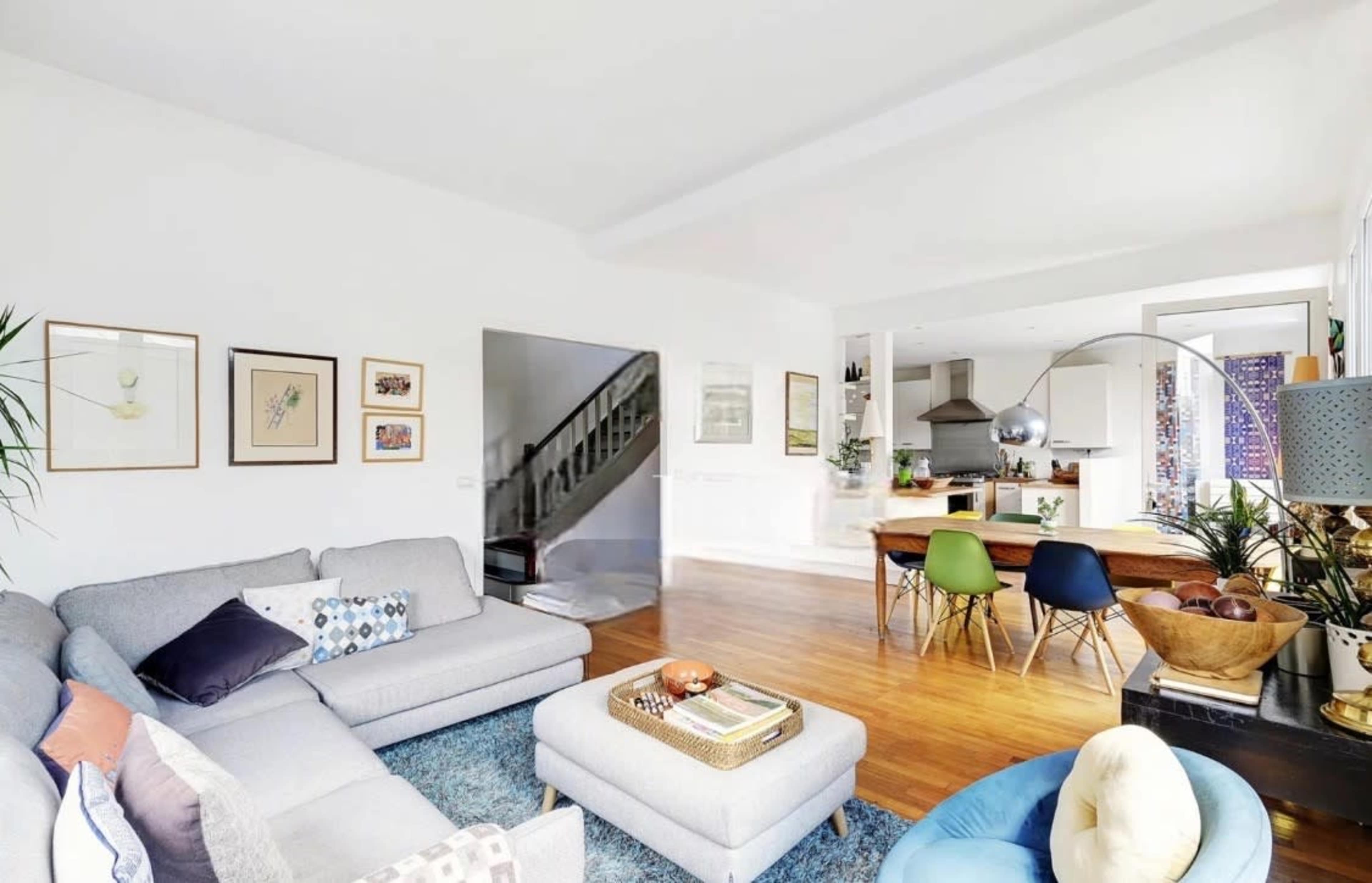 The image shows a bright living area featuring a gray sectional sofa, a coffee table, and a dining area with a wooden table surrounded by colorful chairs.