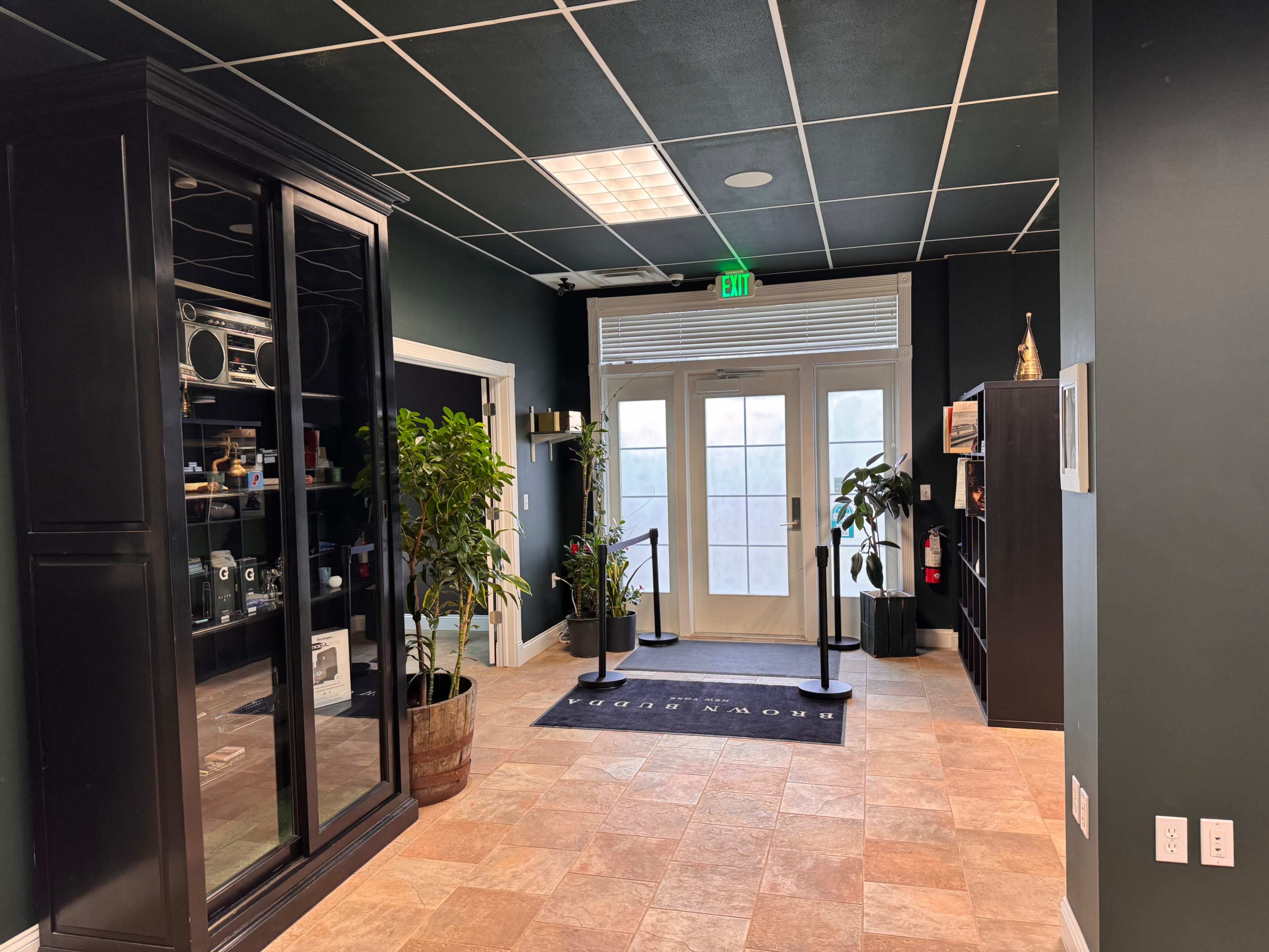 The image shows a lobby area with a glass display cabinet, potted plants, and doors leading outside, flanked by black shelving units.