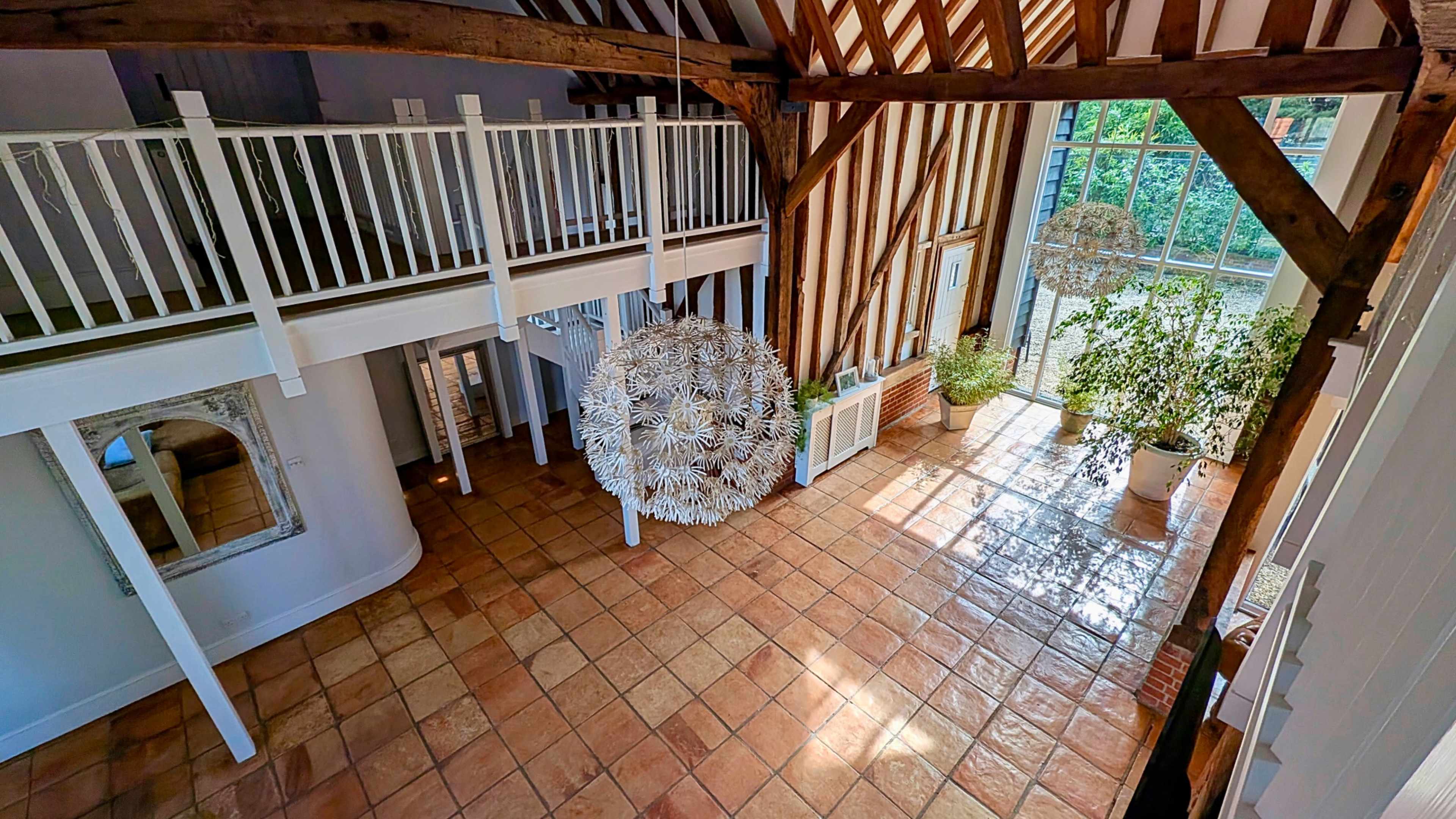 The image shows a spacious indoor area with a tiled floor, large windows, and wooden beams, featuring a decorative spherical light fixture and potted plants.