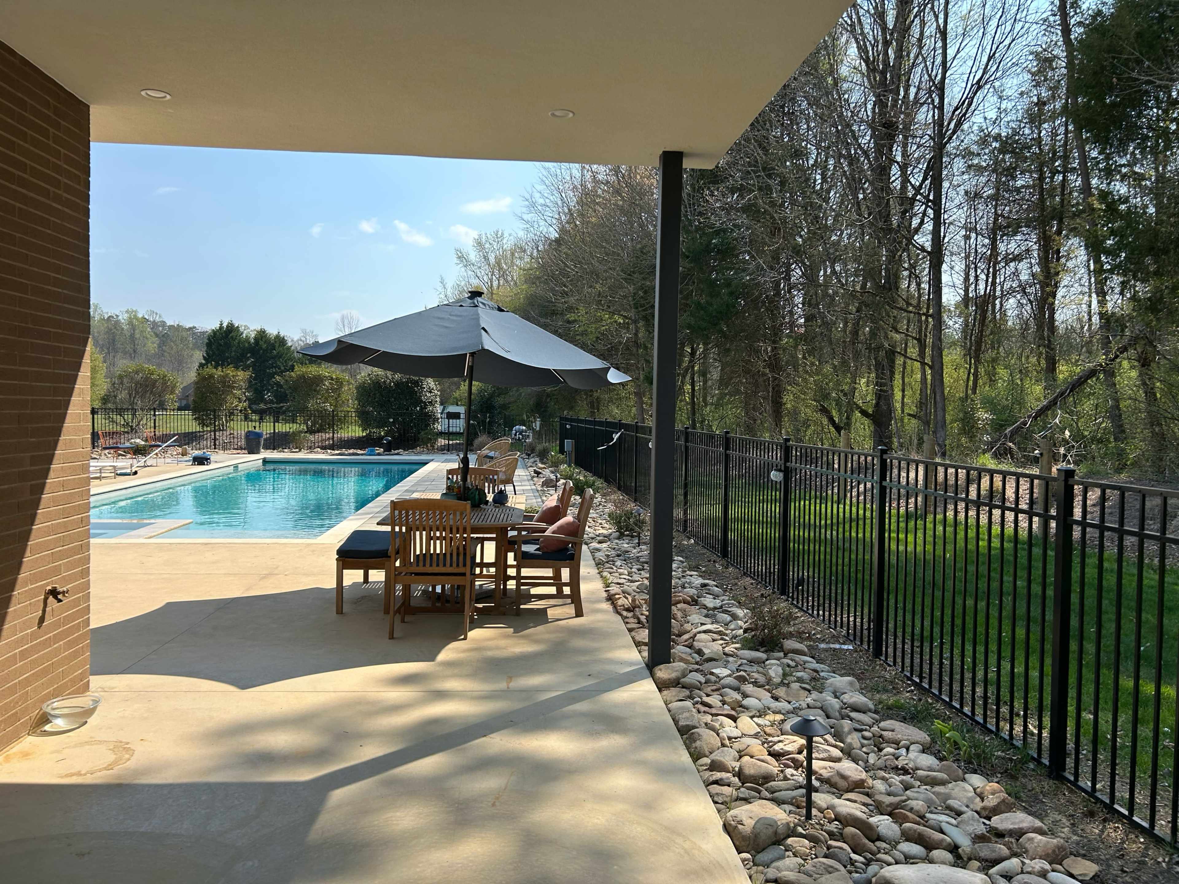 The image shows a patio area next to a swimming pool, with outdoor furniture and an umbrella, surrounded by a fenced backyard and trees.