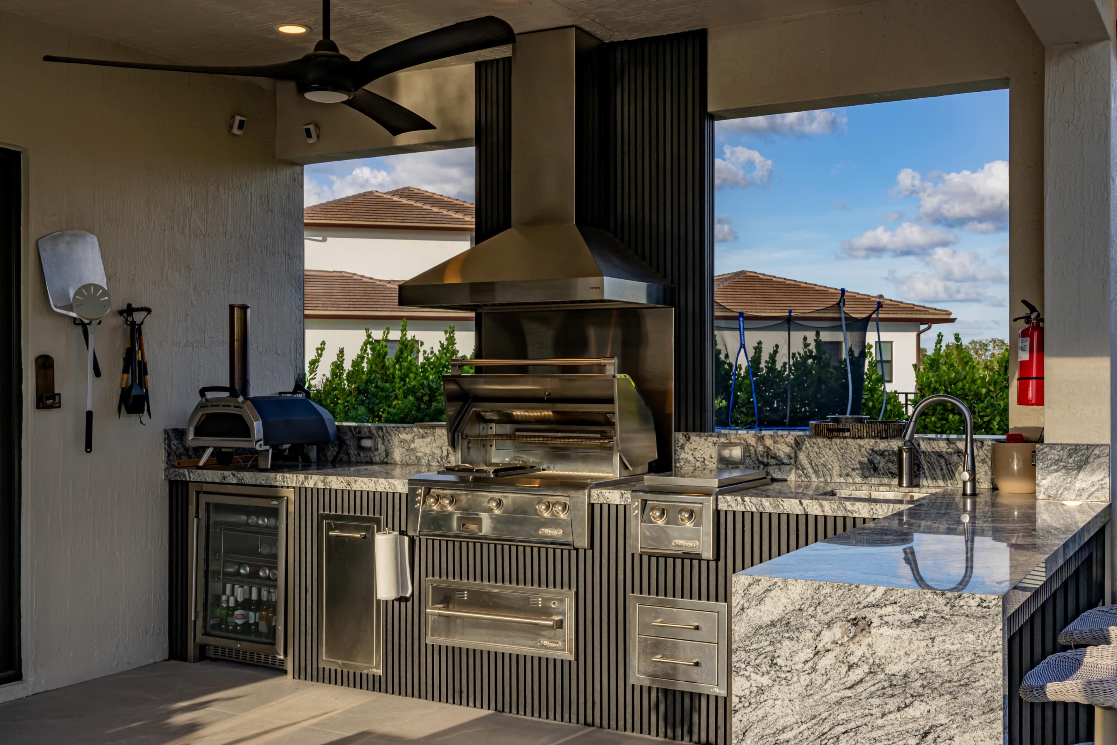 The image shows an outdoor kitchen featuring a stainless steel grill, a sink, and a bar with granite countertops, set against a backdrop of trees and a clear sky.