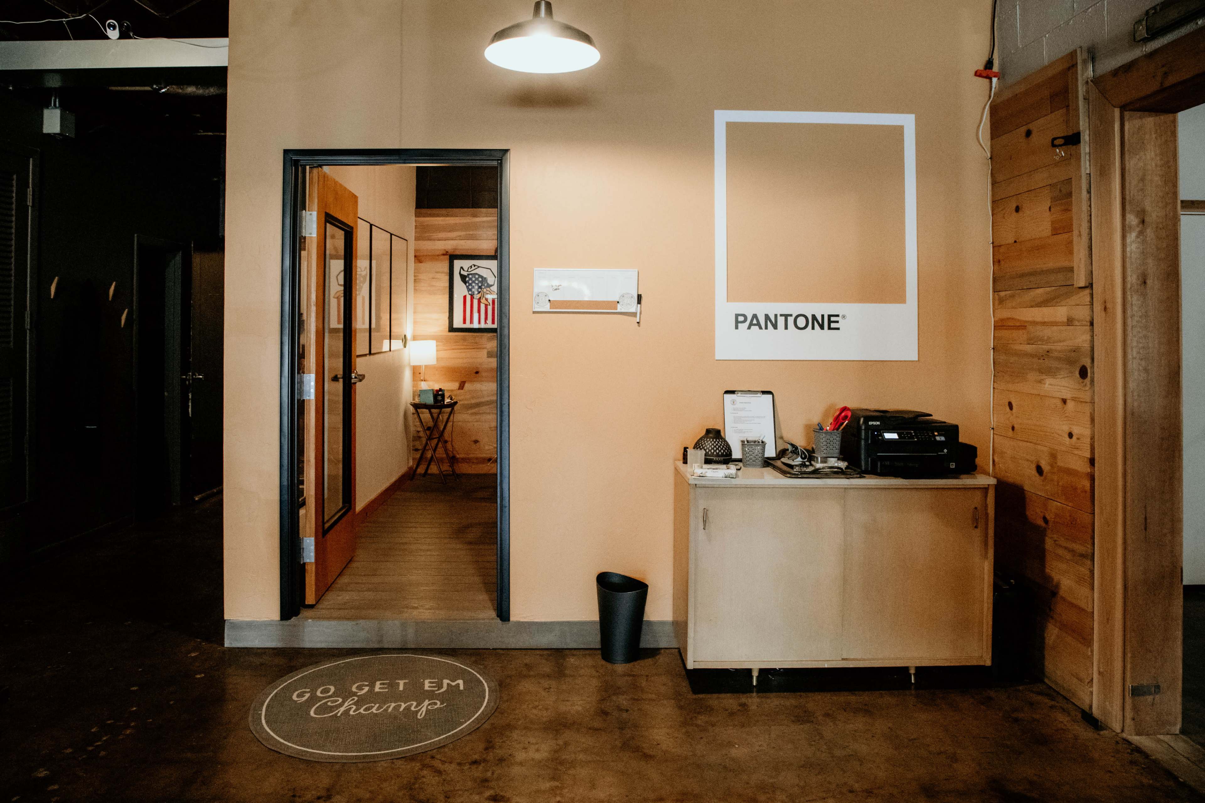An interior space with a reception area featuring a desk, printer, and wall art, alongside an open doorway leading to another room.