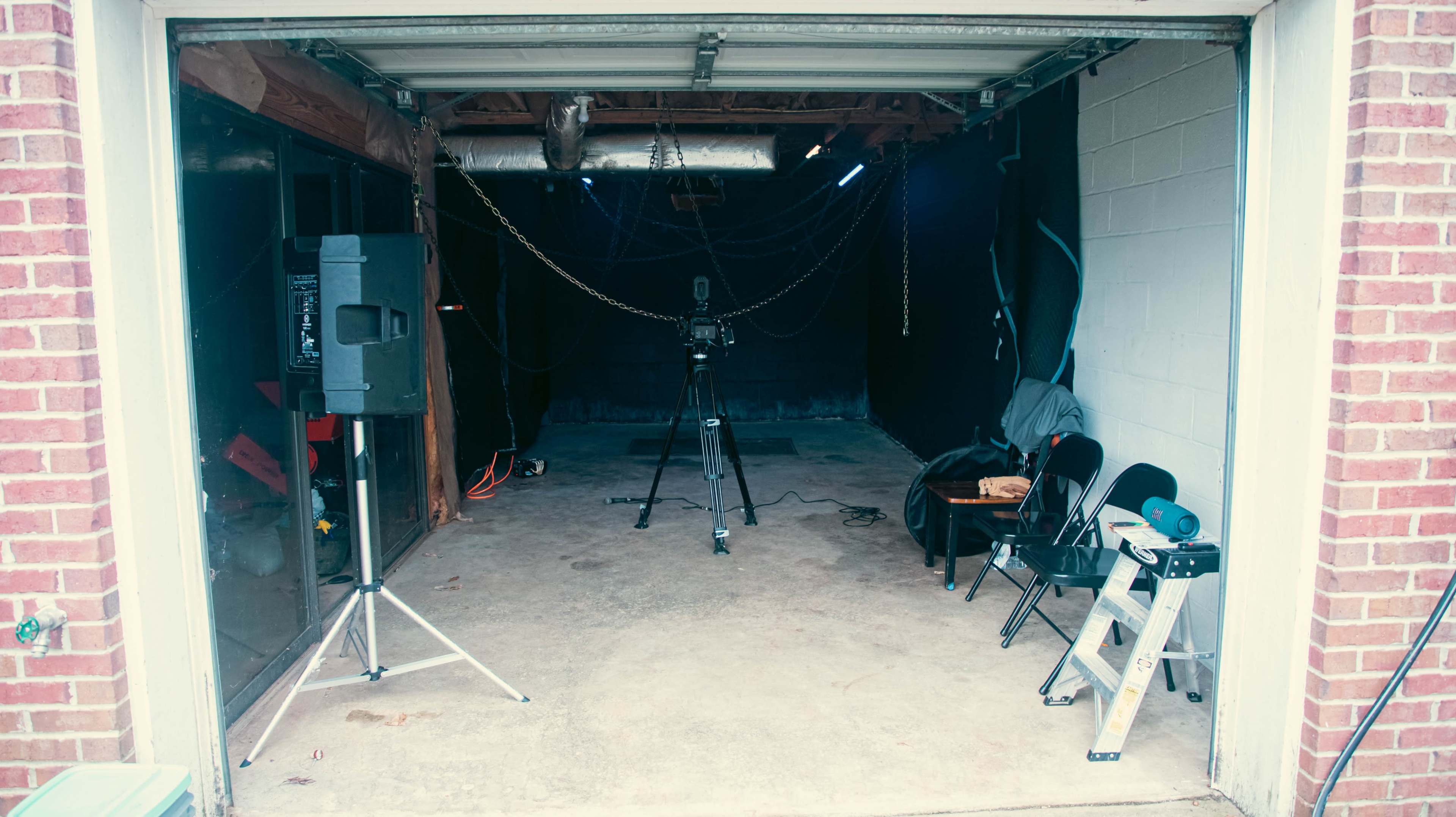 The image shows a partially finished garage space with a camera on a tripod, a speaker, a ladder, and several chairs arranged inside.