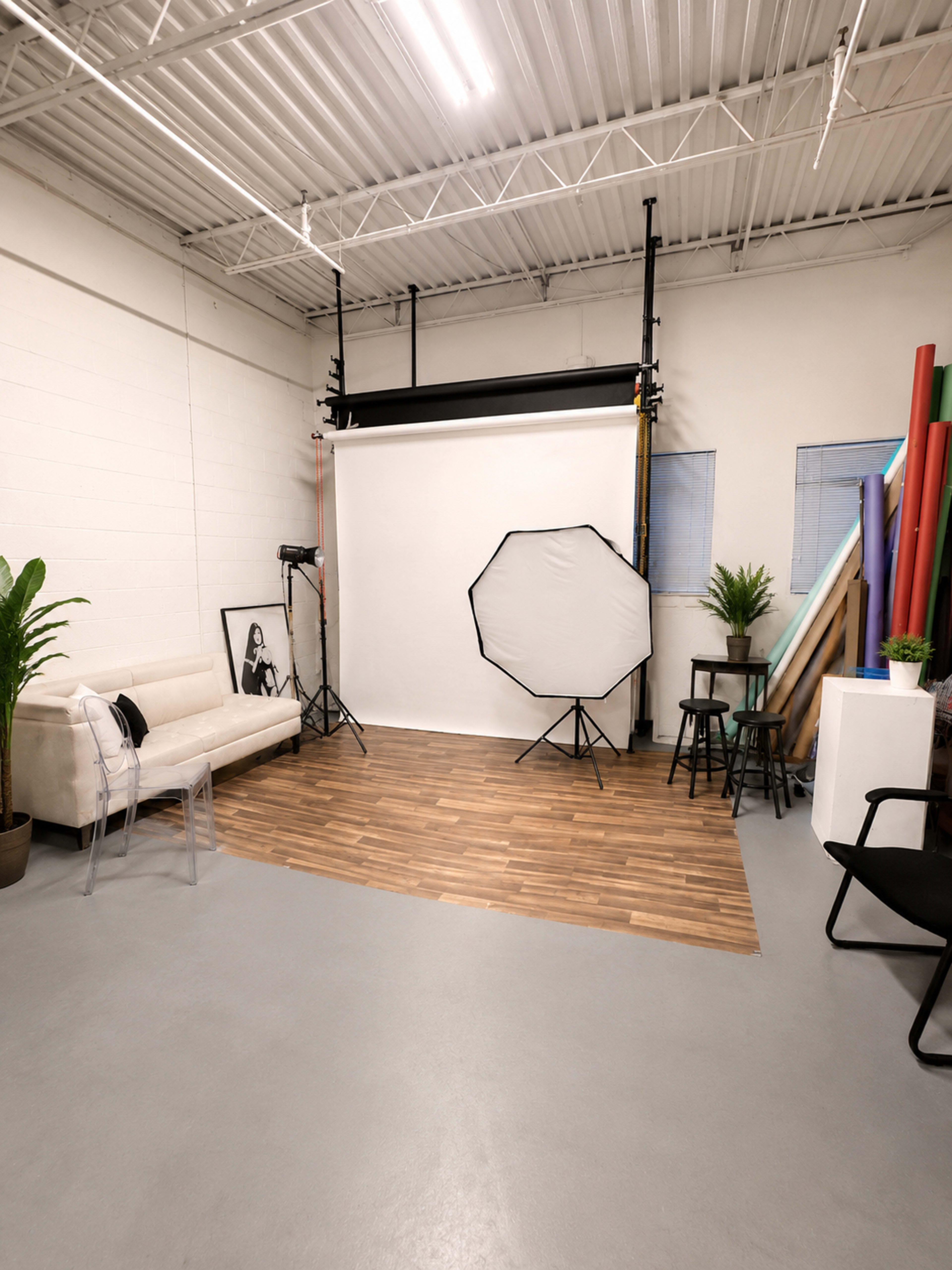 The image shows a photography studio with a white backdrop, a sofa, a chair, and various props and backgrounds arranged along the walls.