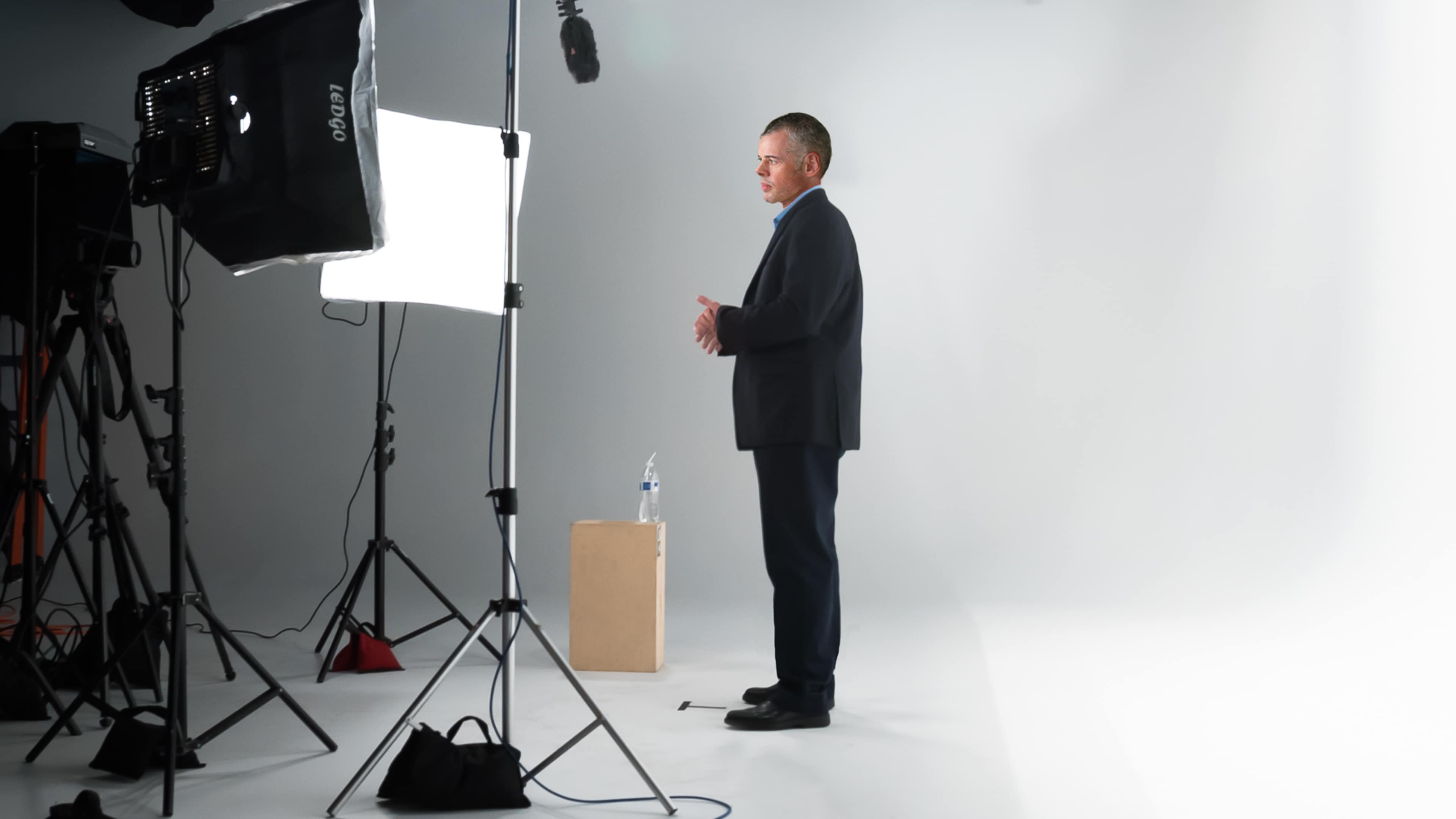 A man in a suit stands beside a microphone and a box on a plain white backdrop, with lighting equipment set up around him.