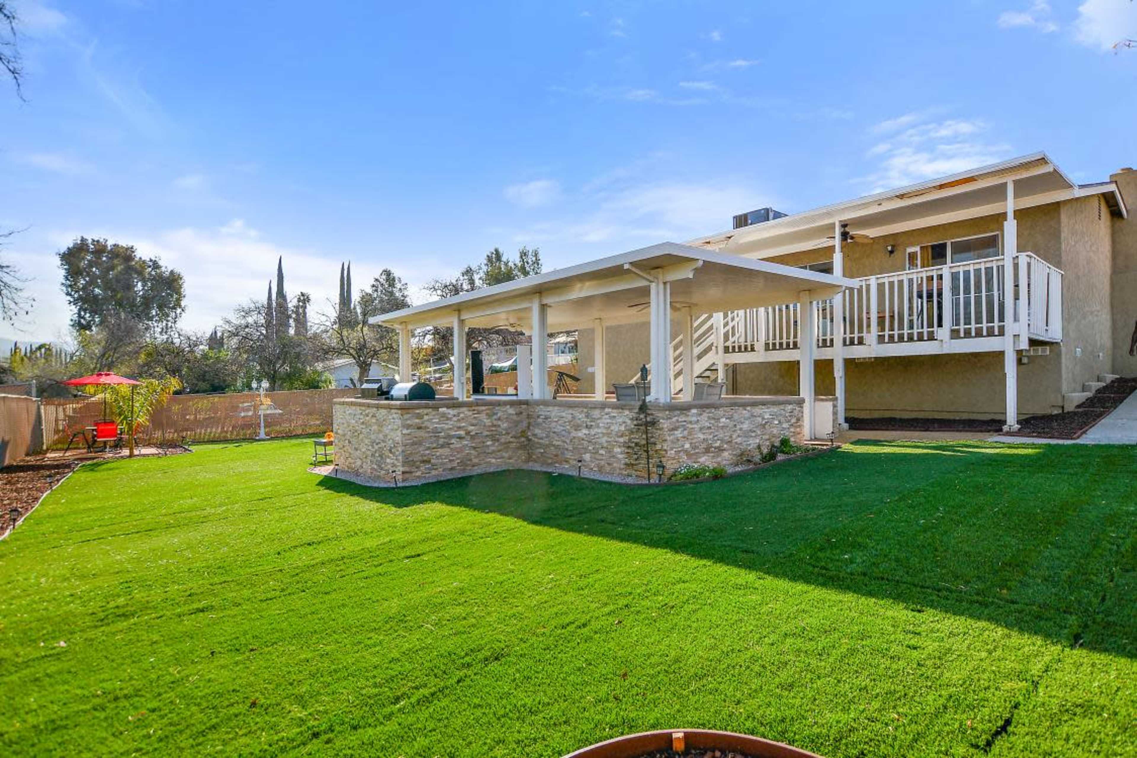 A well-maintained backyard features a grassy lawn, a covered patio with stone accents, and a single-story house in the background.