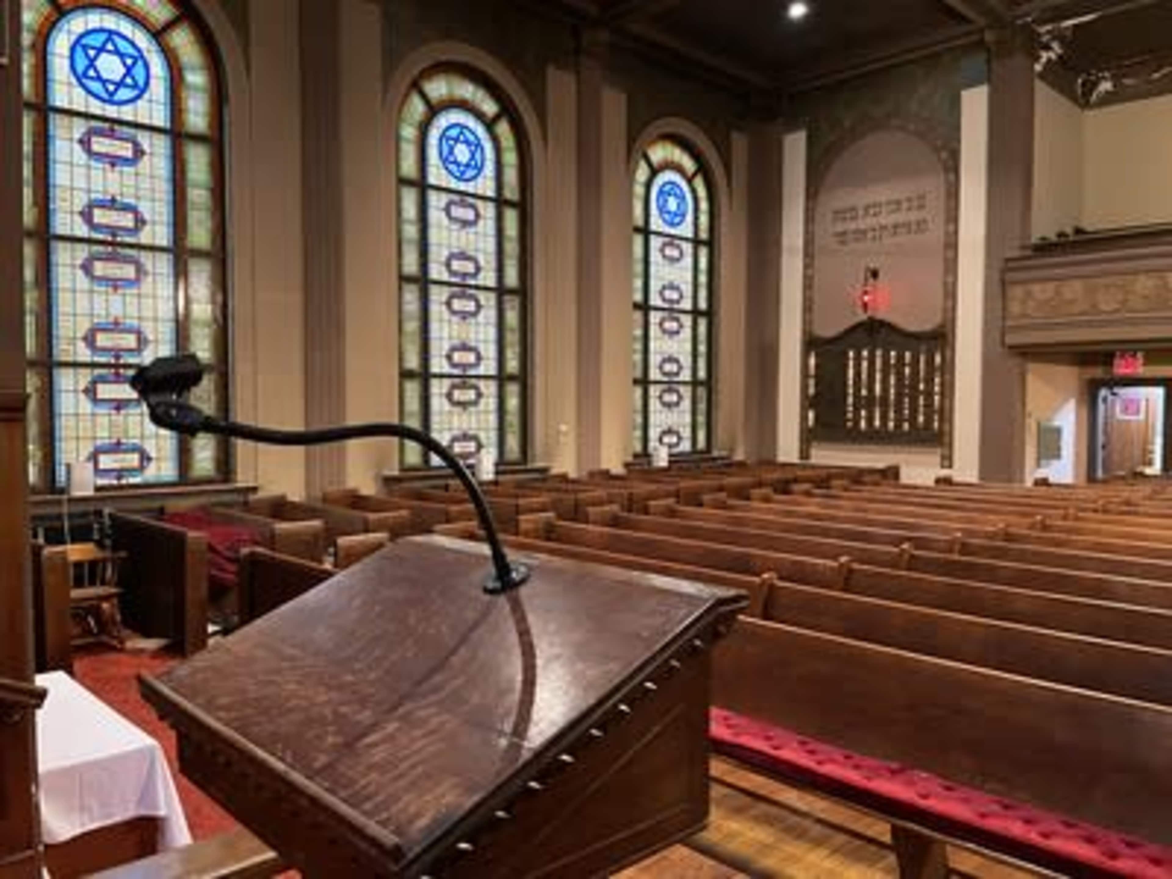The interior of a synagogue with a wooden podium in the foreground, surrounded by rows of wooden benches and stained glass windows featuring Star of David symbols.
