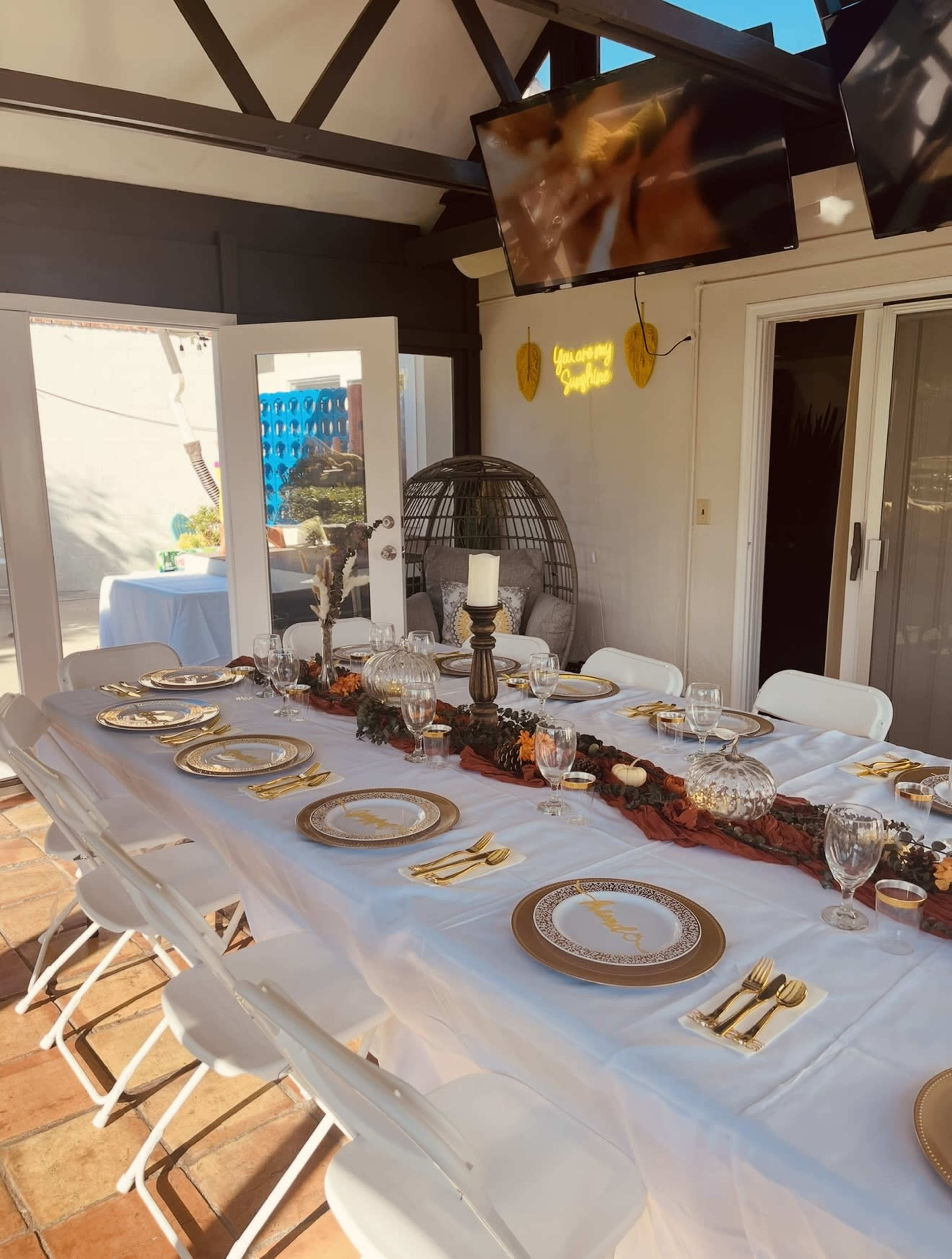 A long dining table is elegantly set with gold-rimmed plates, glassware, and utensils under a shaded outdoor area featuring two mounted televisions.