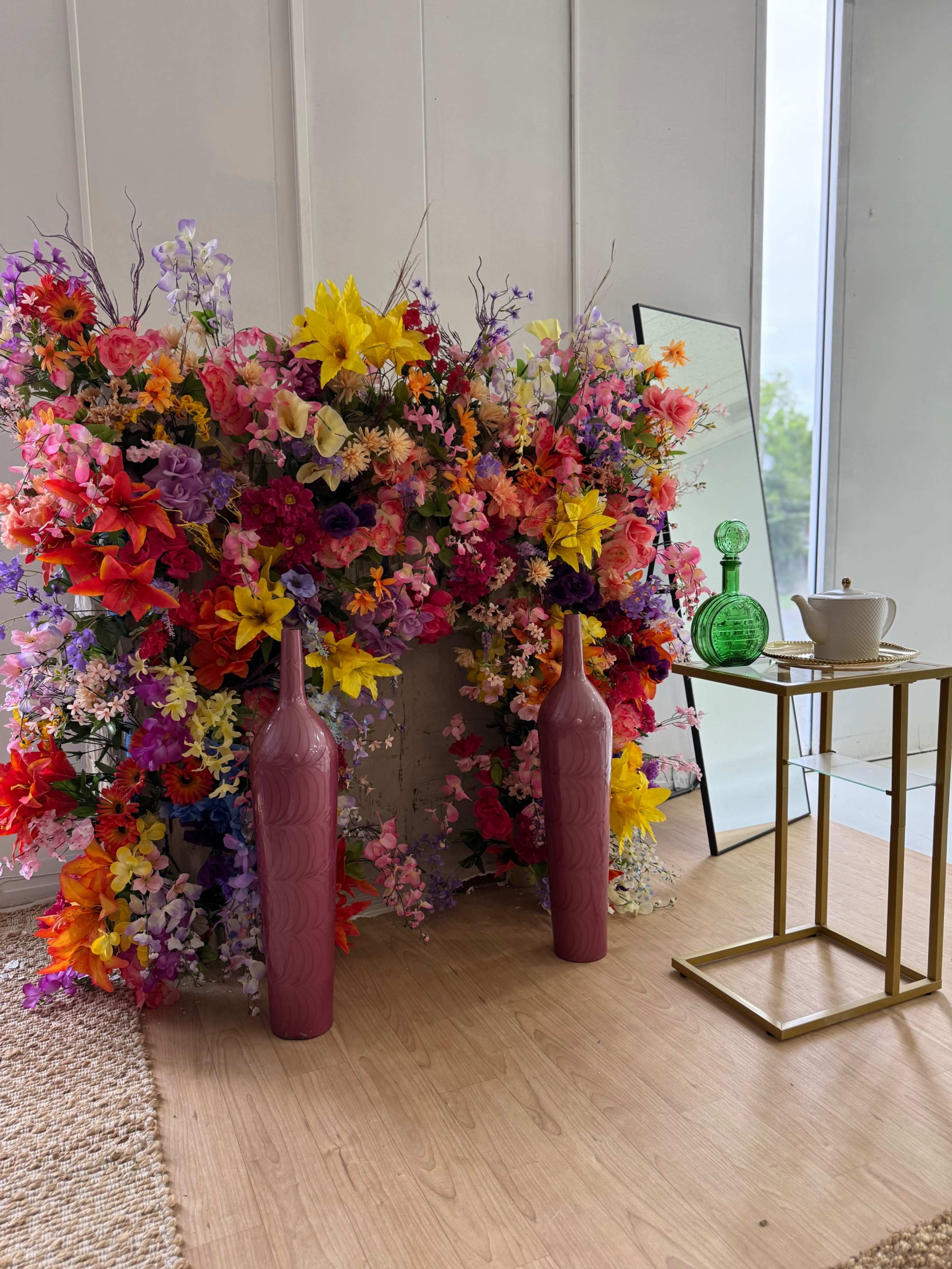 A vibrant floral arrangement composed of various colorful flowers stands behind two tall vases, alongside a small table with a green decorative bottle and a white cup.