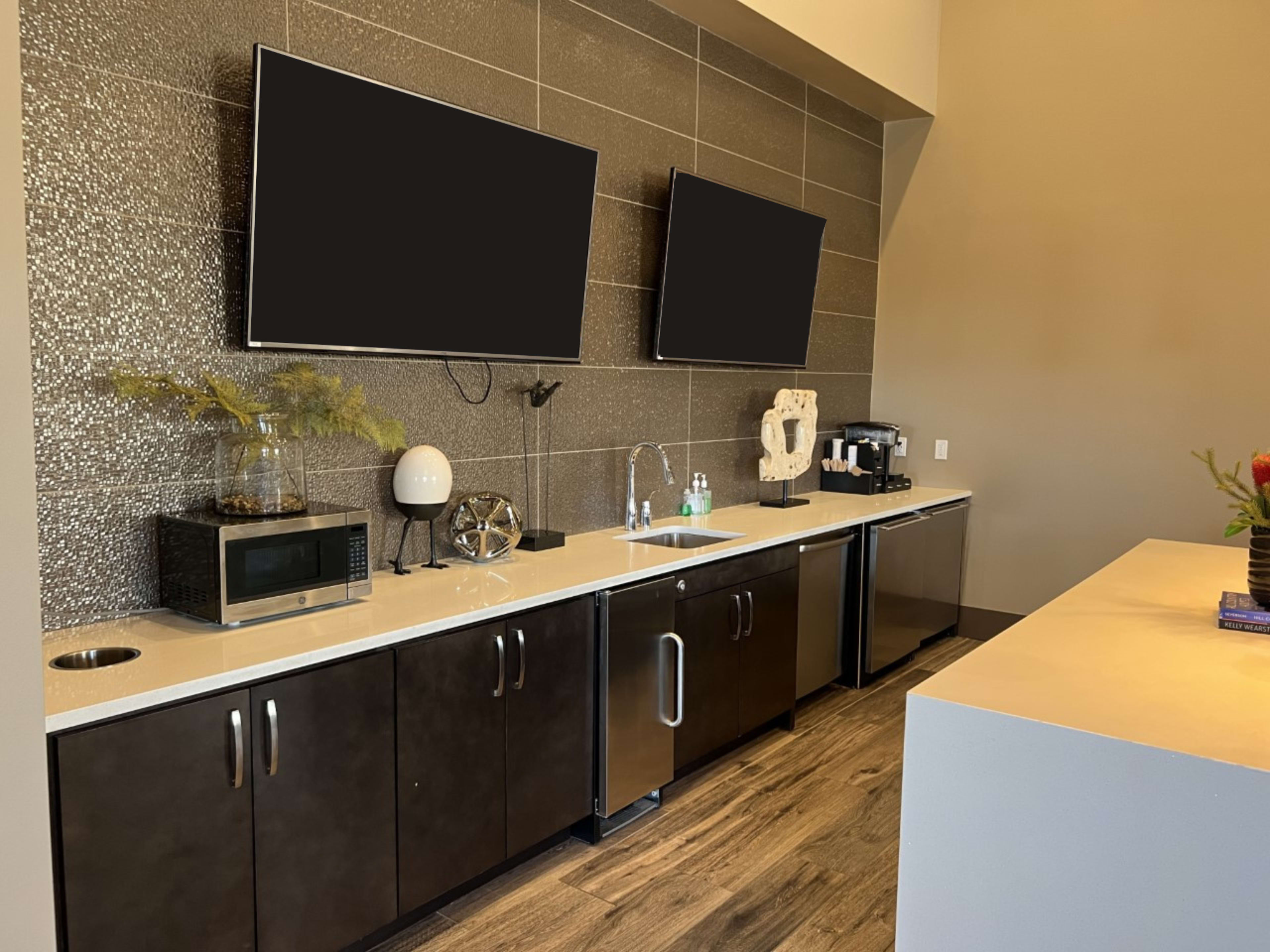 A modern kitchenette with two wall-mounted televisions above a counter featuring a microwave, coffee maker, and various decorative items.