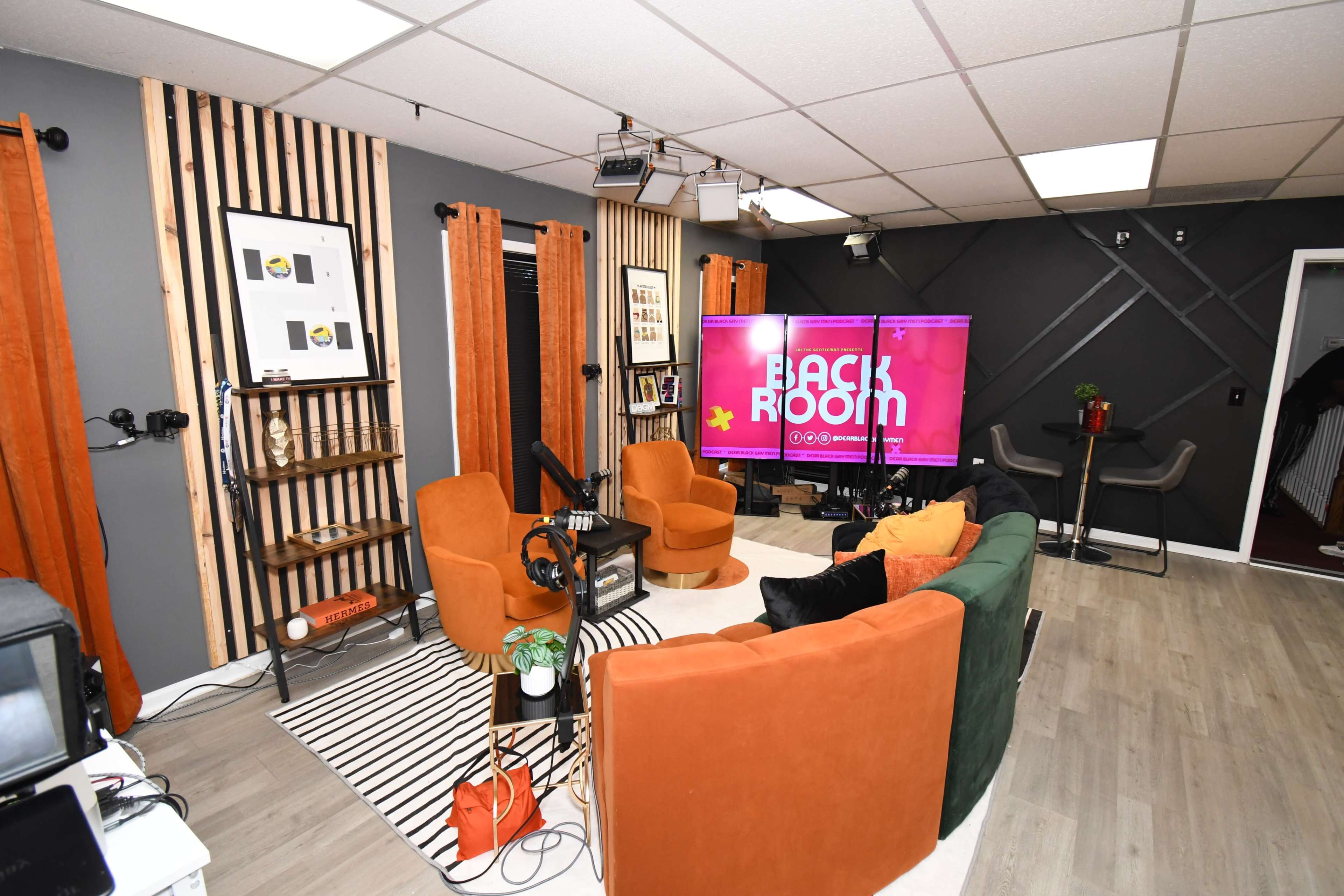 A modern studio space features orange and green furniture, a large screen displaying "BACK ROOM," and decorative elements against a wood-paneled wall.
