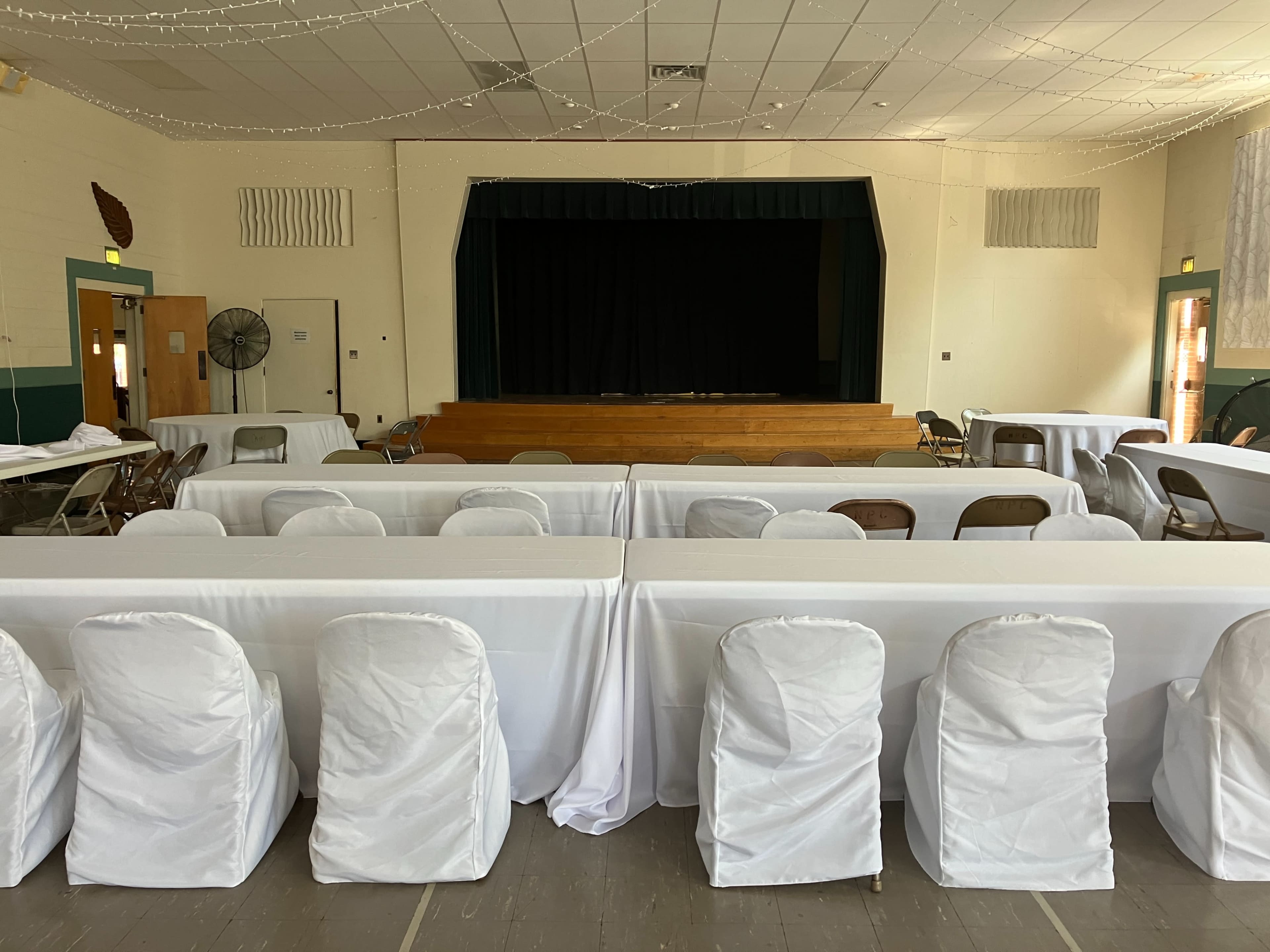A spacious indoor event venue with rows of tables covered in white tablecloths and chairs wrapped in white coverings, facing a stage with a black curtain.