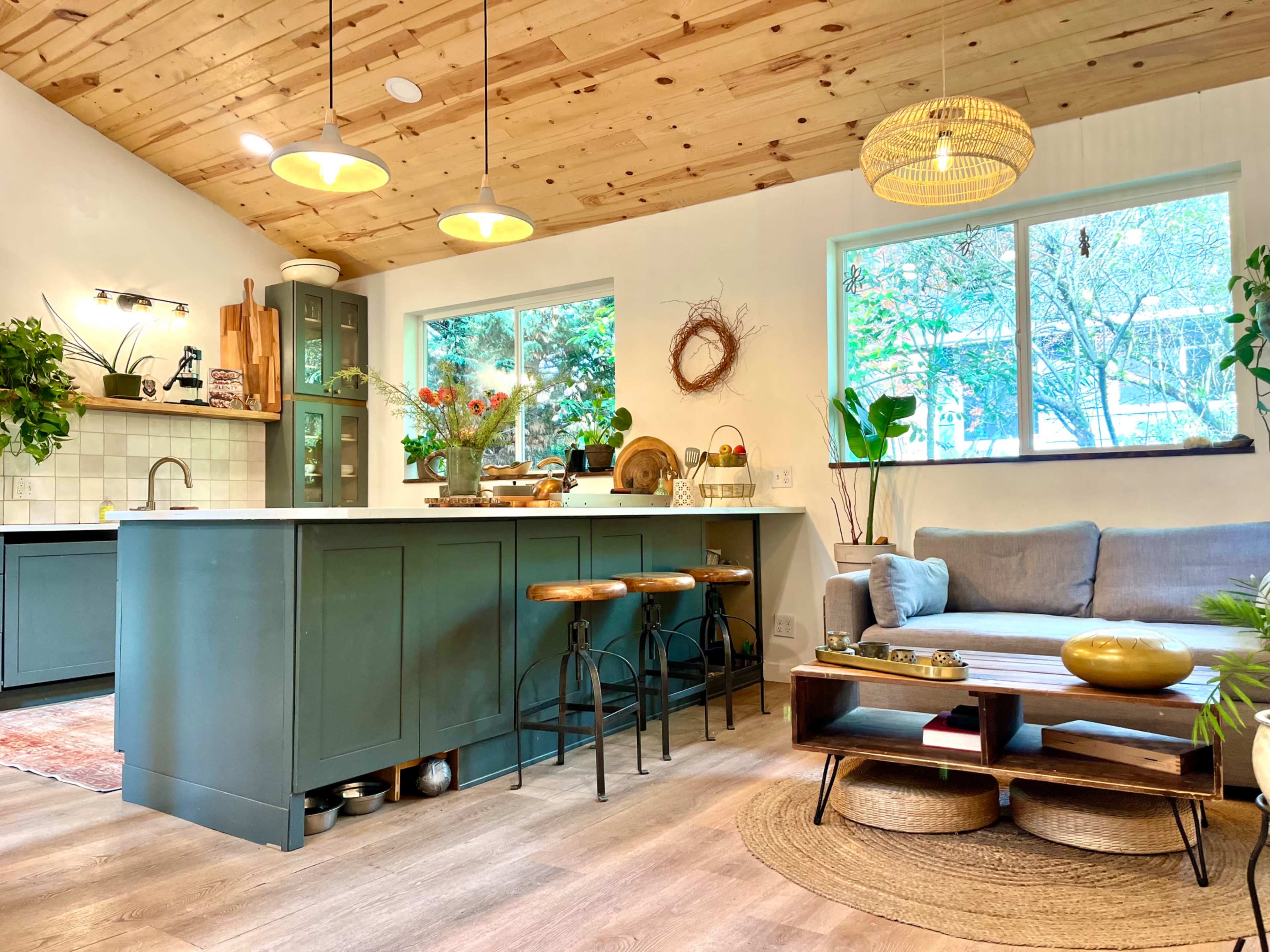 The image features a modern kitchen with a wooden ceiling, green cabinets, and a cozy seating area with a gray sofa and a wooden coffee table.
