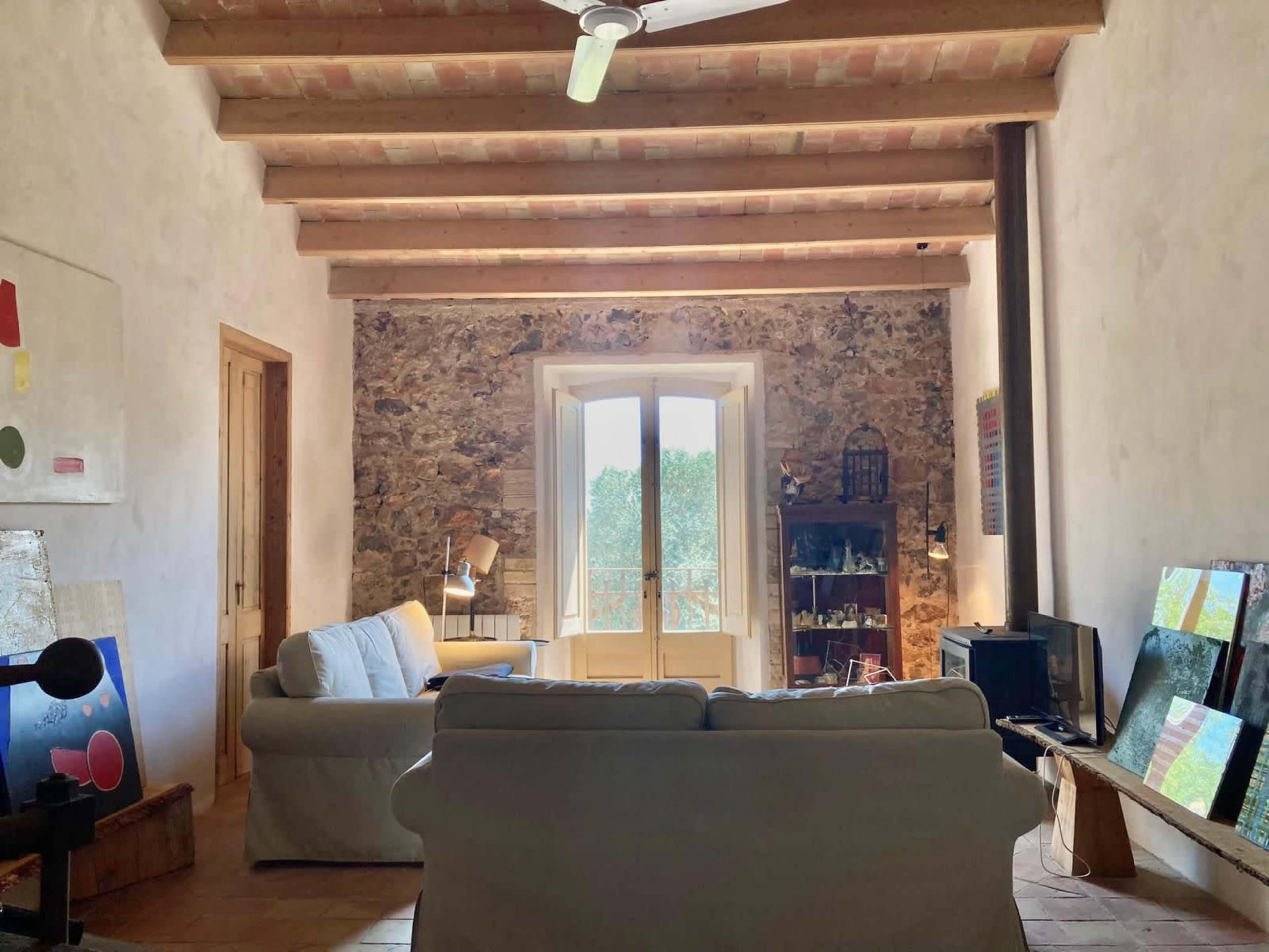 A living room with a white sofa, a stone wall, wooden ceiling beams, and a window that opens to a view of greenery outside.