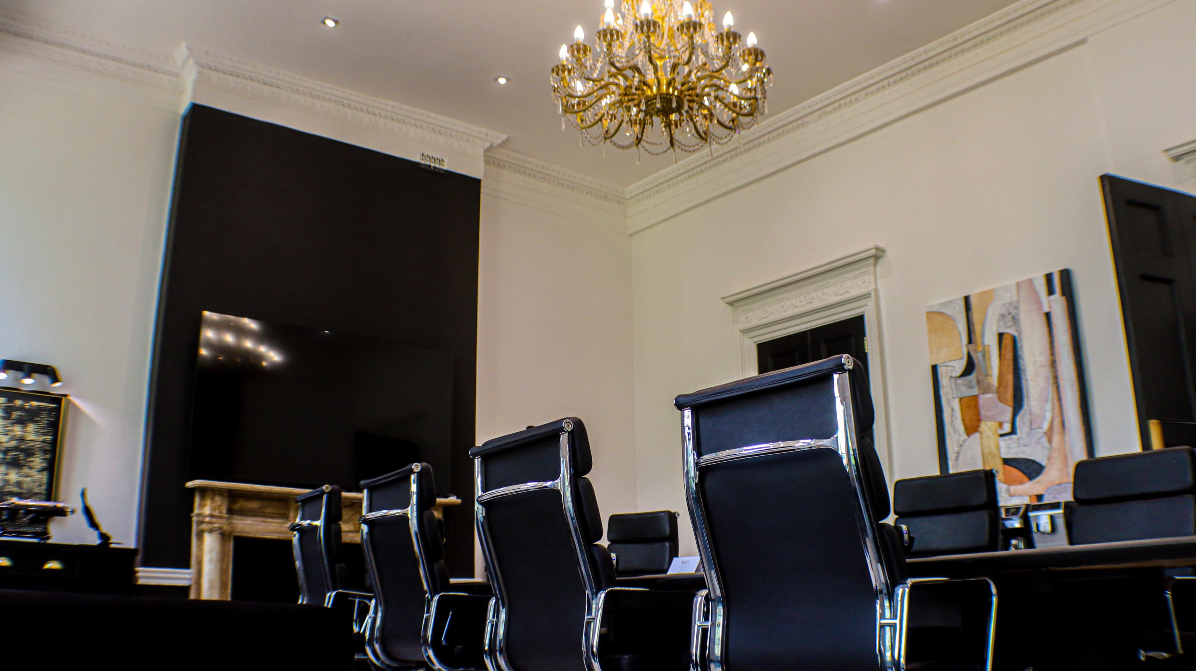 A modern conference room features a long table surrounded by black leather chairs, with a large chandelier overhead and an artwork displayed on the wall.