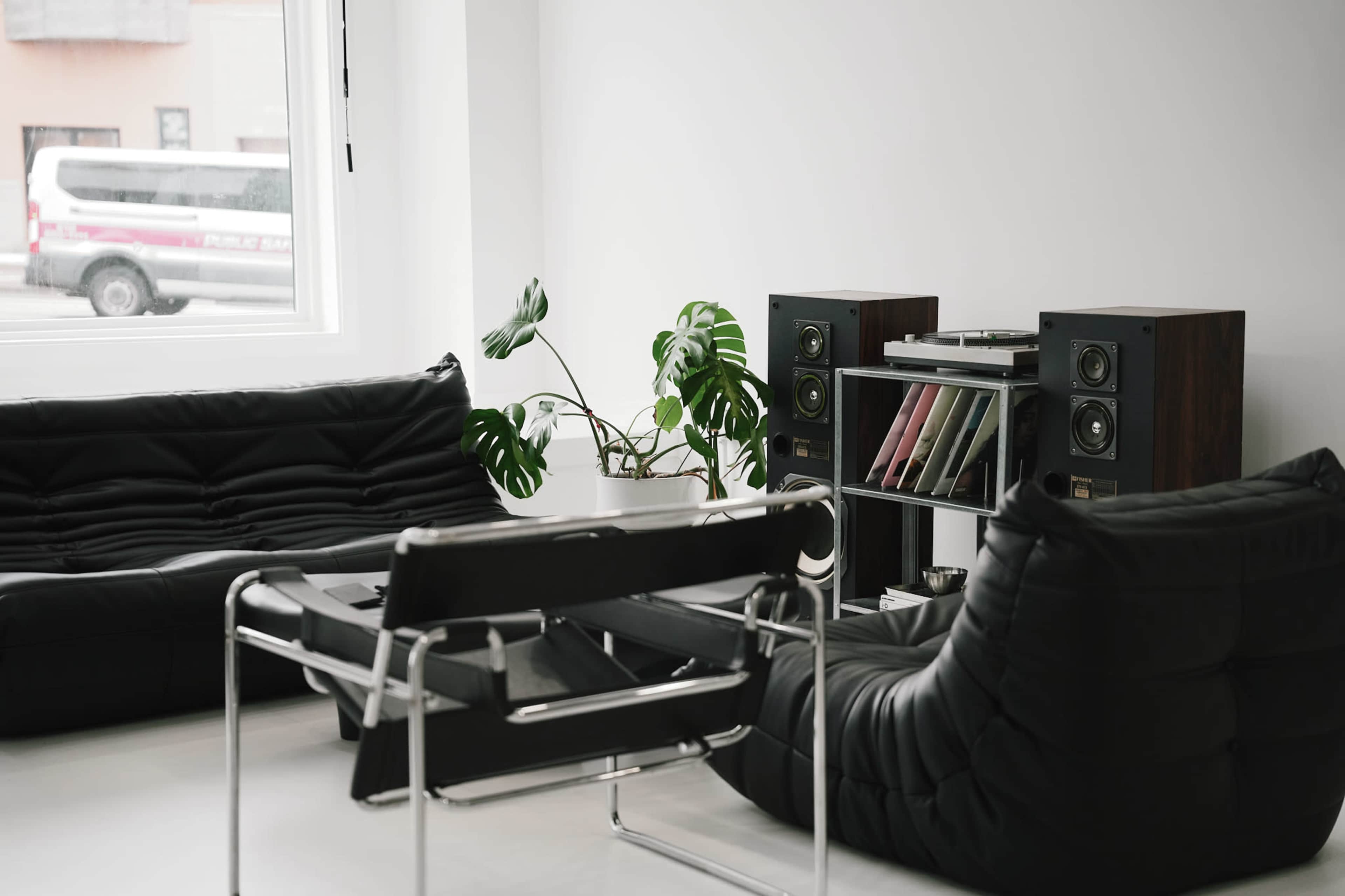 The image shows a modern living room with black furniture, including a pair of cushioned sofas and a sleek chair, alongside a record player and speakers on a shelf.