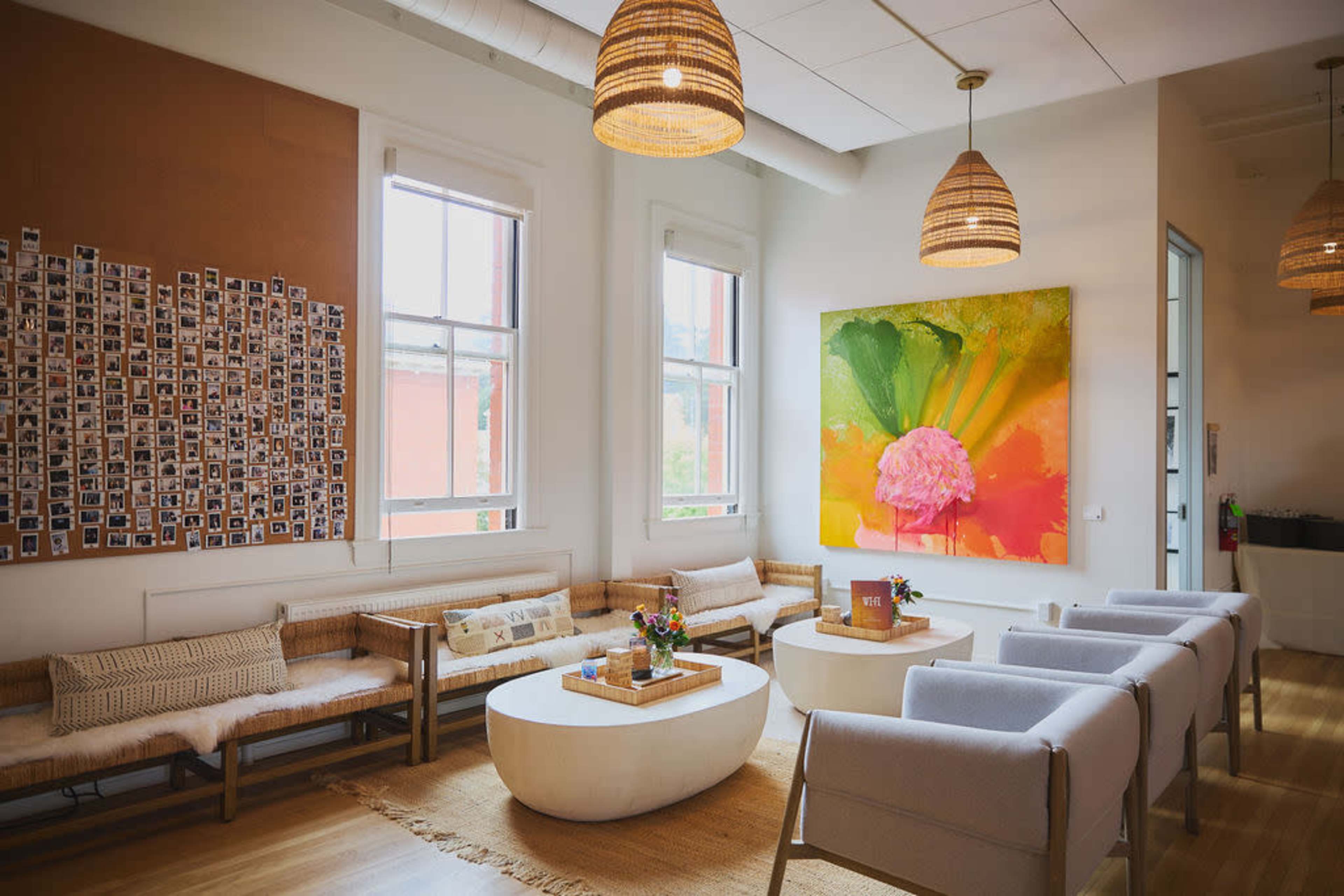 The image shows a modern lounge area with wooden benches, plush seating, a large colorful painting on the wall, and a display of small photographs.