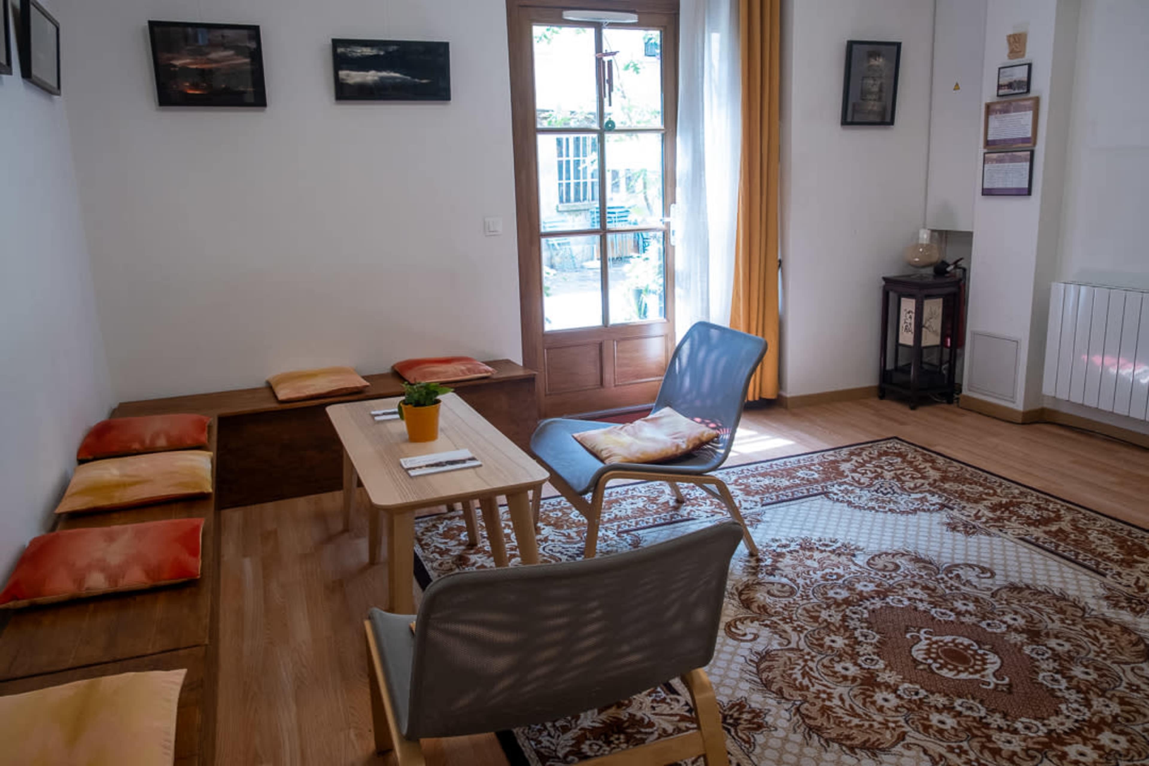 The image shows a cozy room with a wooden table and chairs, a patterned area rug, and a large window that lets in natural light.