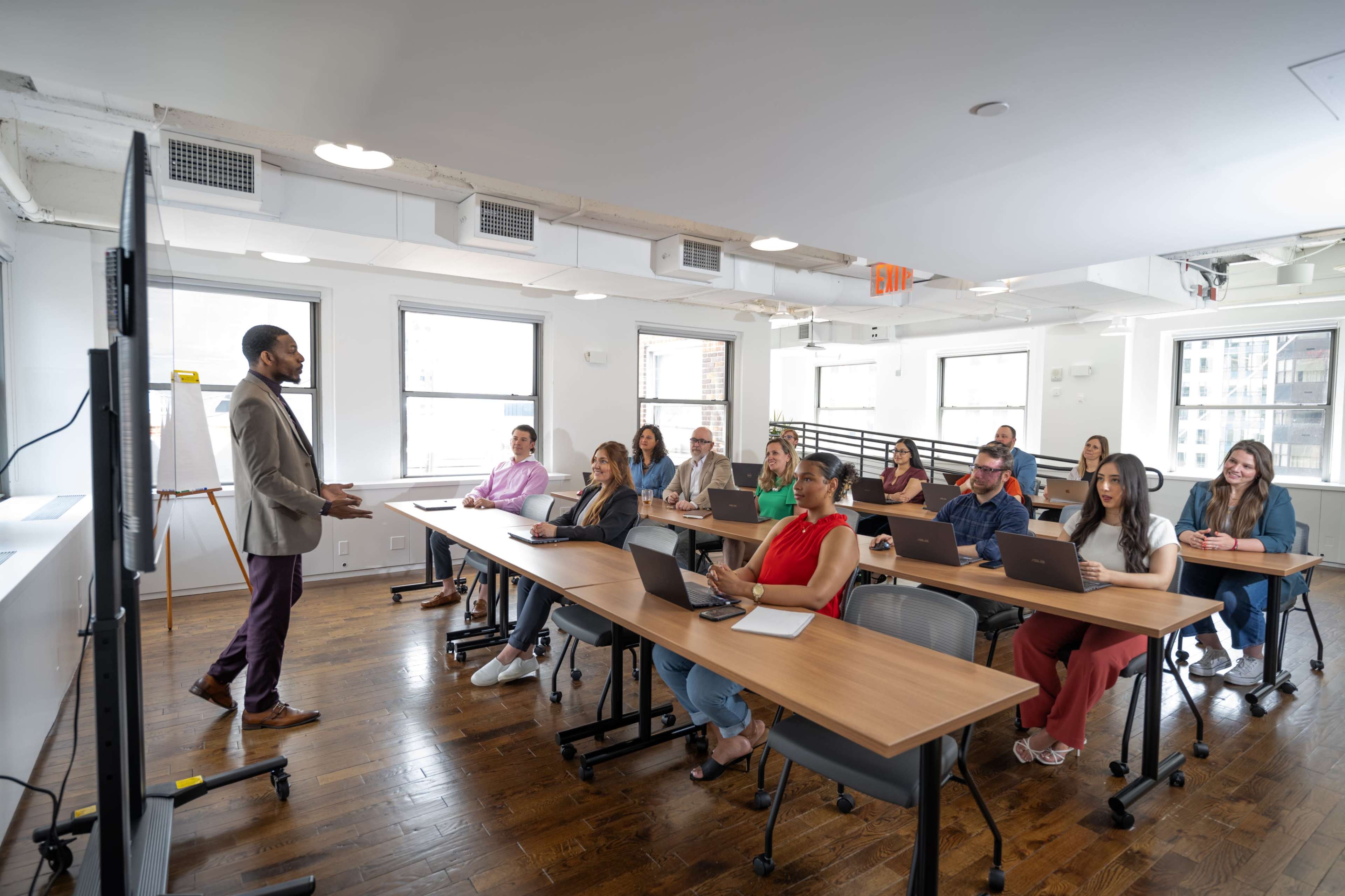 A speaker presents to a seated audience in a modern classroom setting with laptops on their desks.