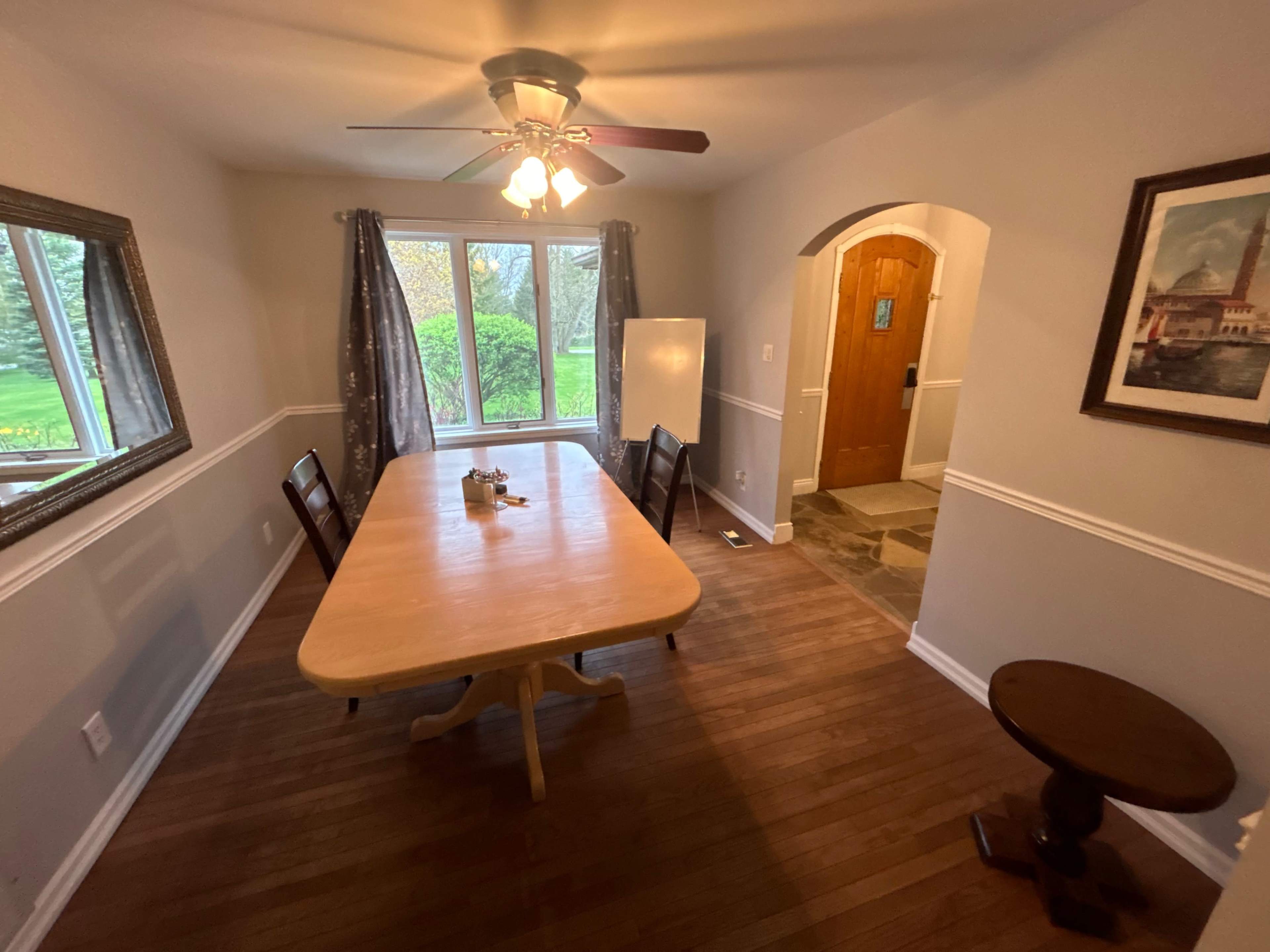 A dining area with a wooden table and chairs, a ceiling fan, and a view of a garden through the windows.