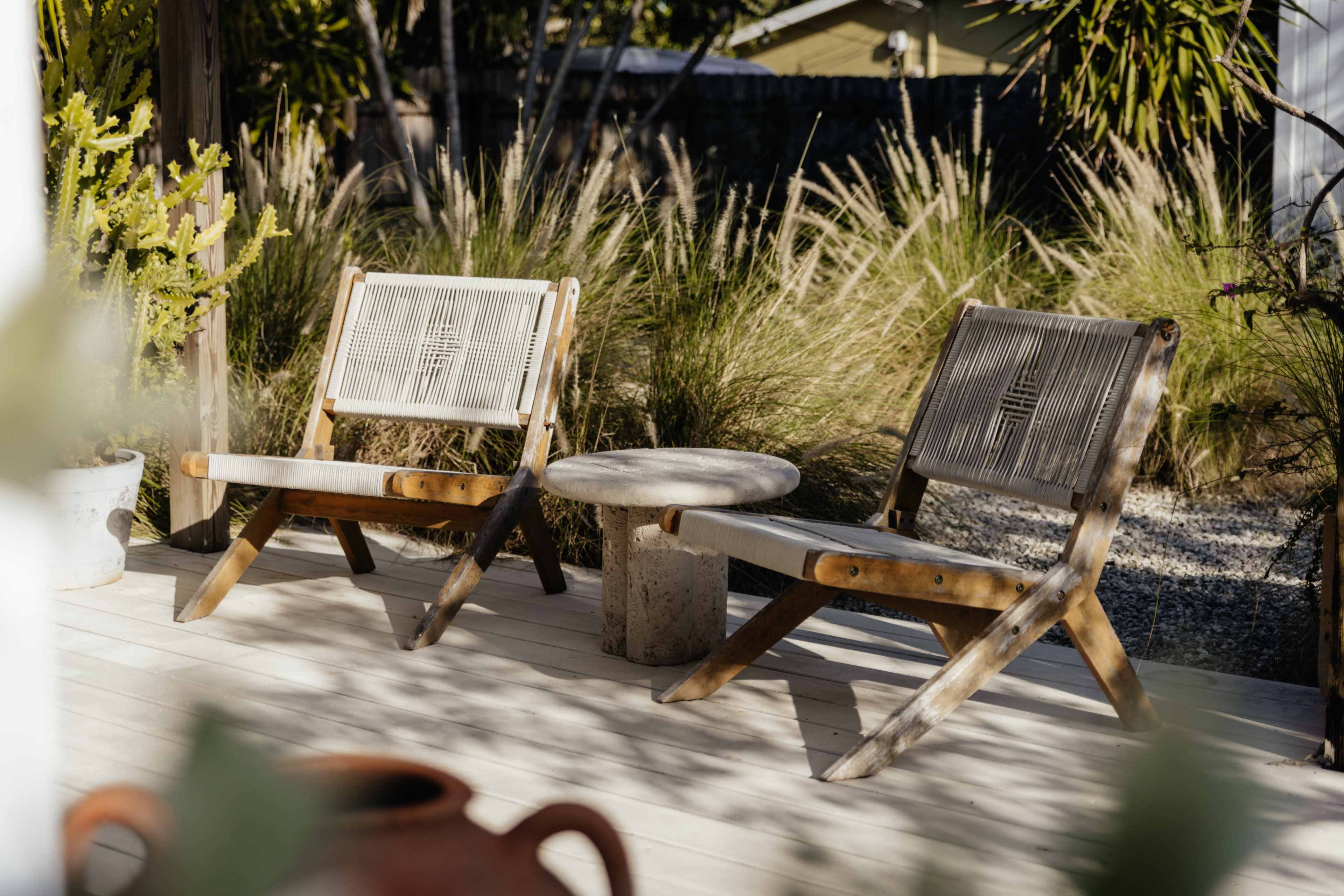 Two wooden chairs with a small round table between them are positioned on a deck surrounded by tall grasses and plants.