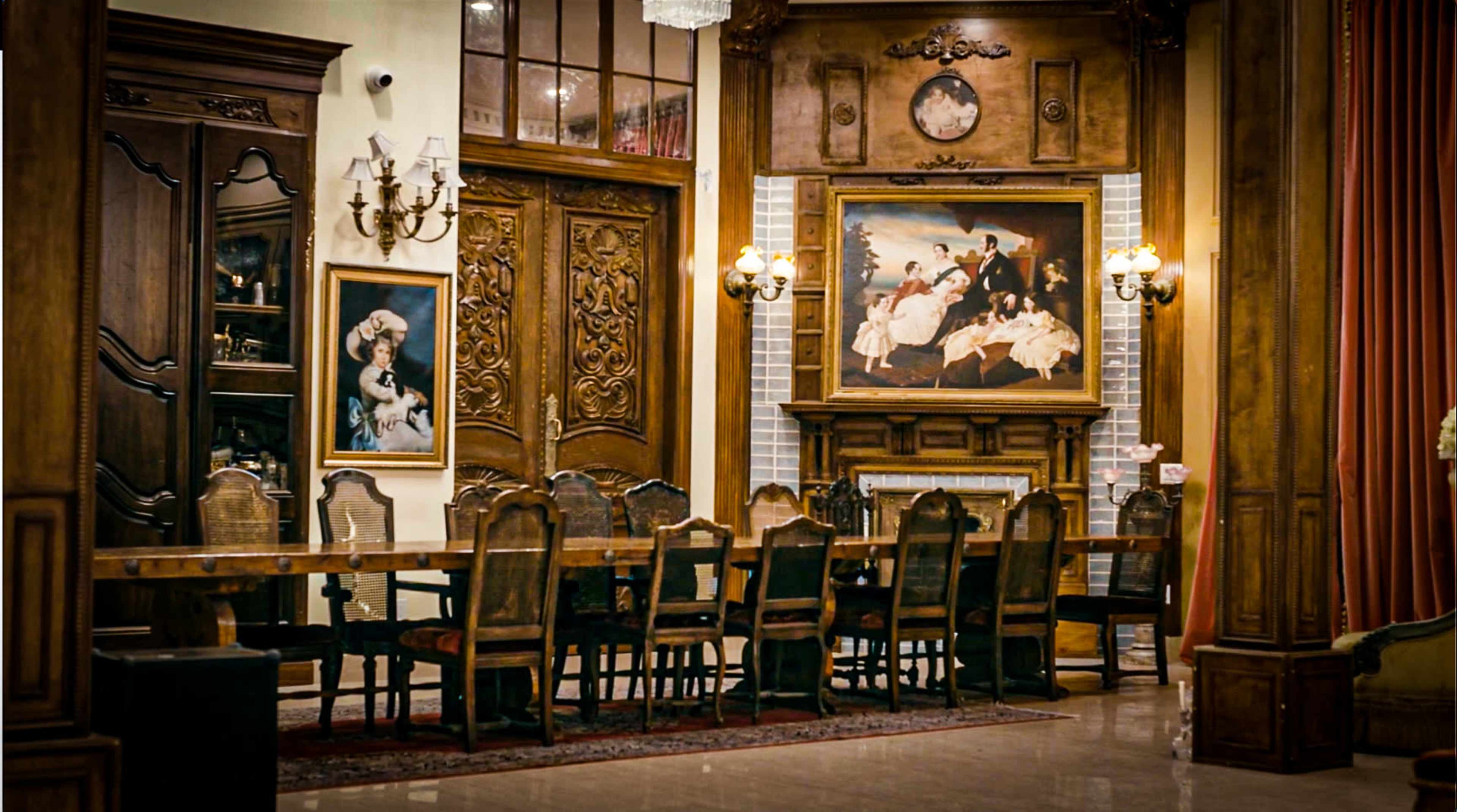 A large dining room features a long wooden table surrounded by ornate chairs, with classical artwork and decorative wall elements.