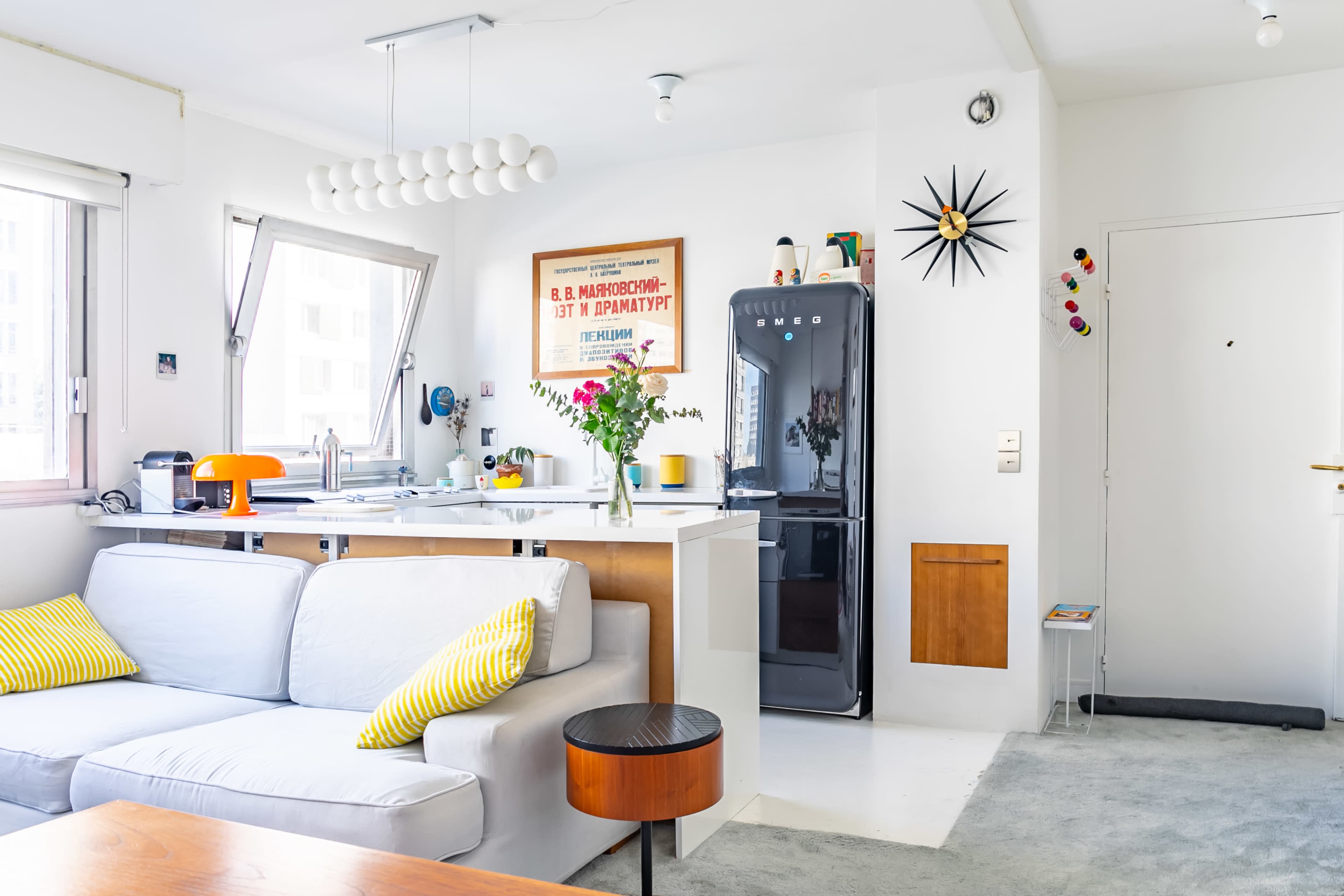 The image shows a modern, bright living space featuring a gray sofa, a kitchen with white cabinetry, a black refrigerator, and a central table with a vase of flowers.