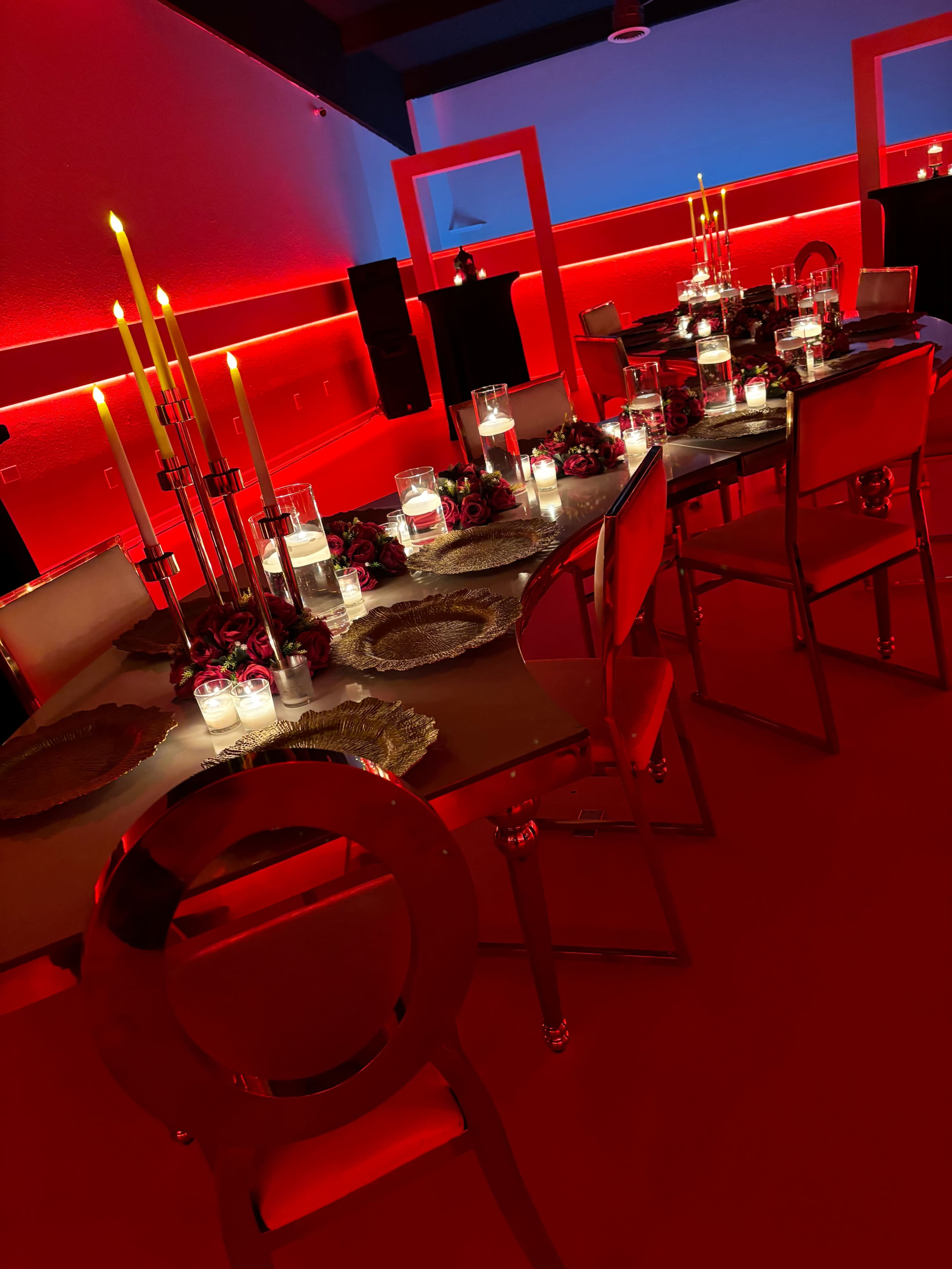 A dining table is elegantly set with candles and floral arrangements, illuminated by red lighting in a modern space.