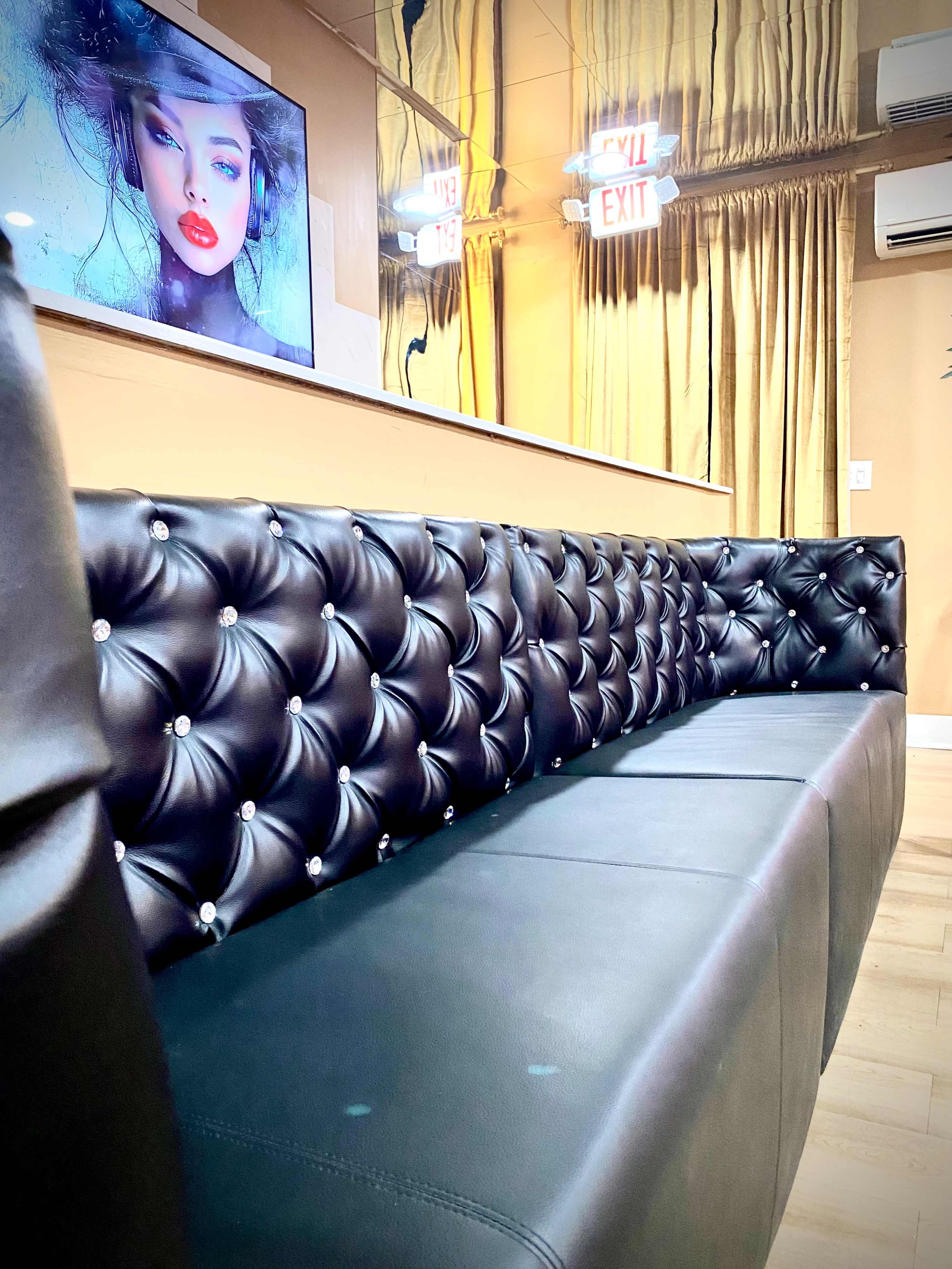 The image shows a black tufted leather bench along a wall, with a large framed portrait of a woman displayed on a screen above it.