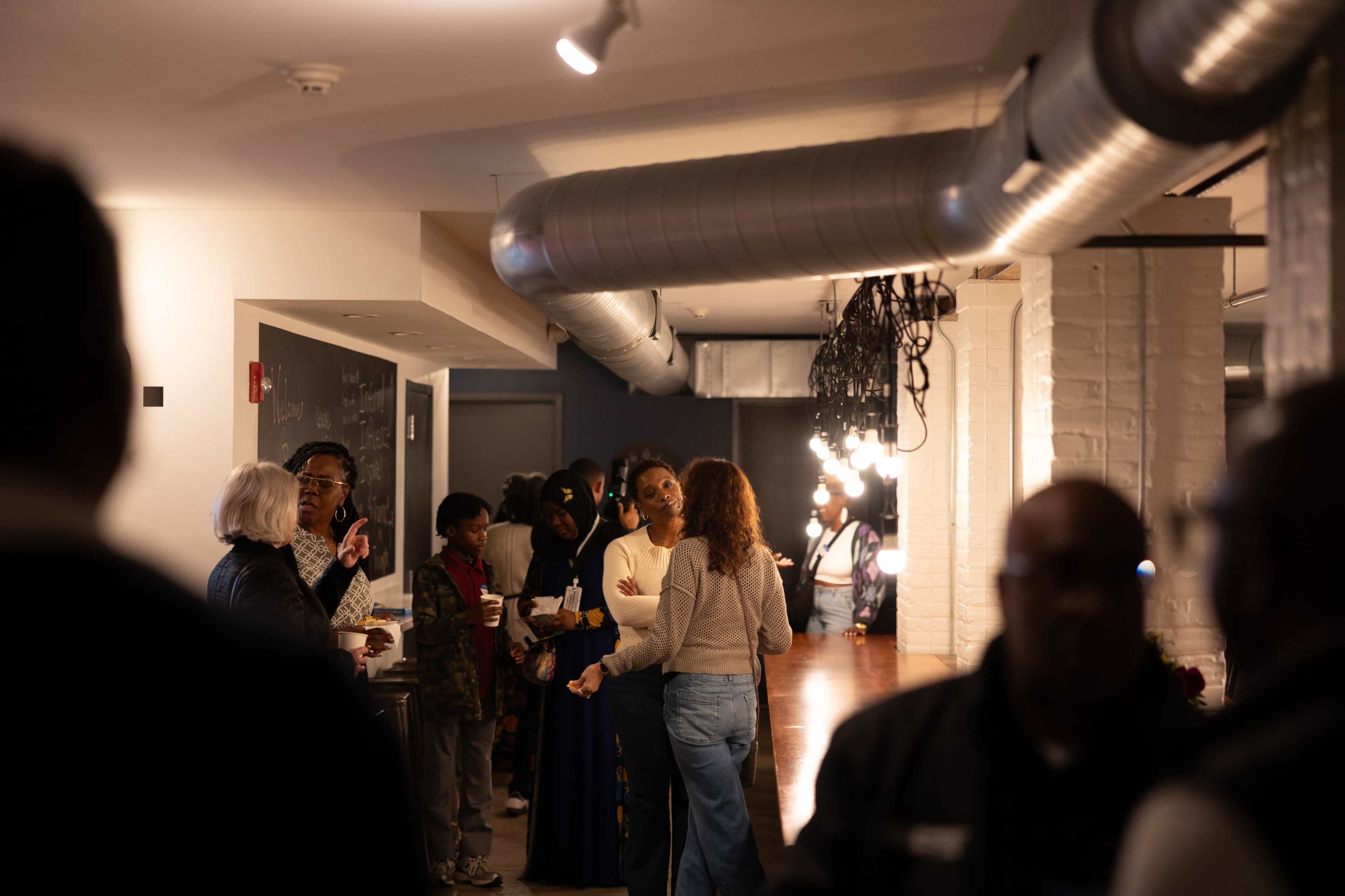 A group of people interacts in a spacious room with exposed ductwork and warm lighting.
