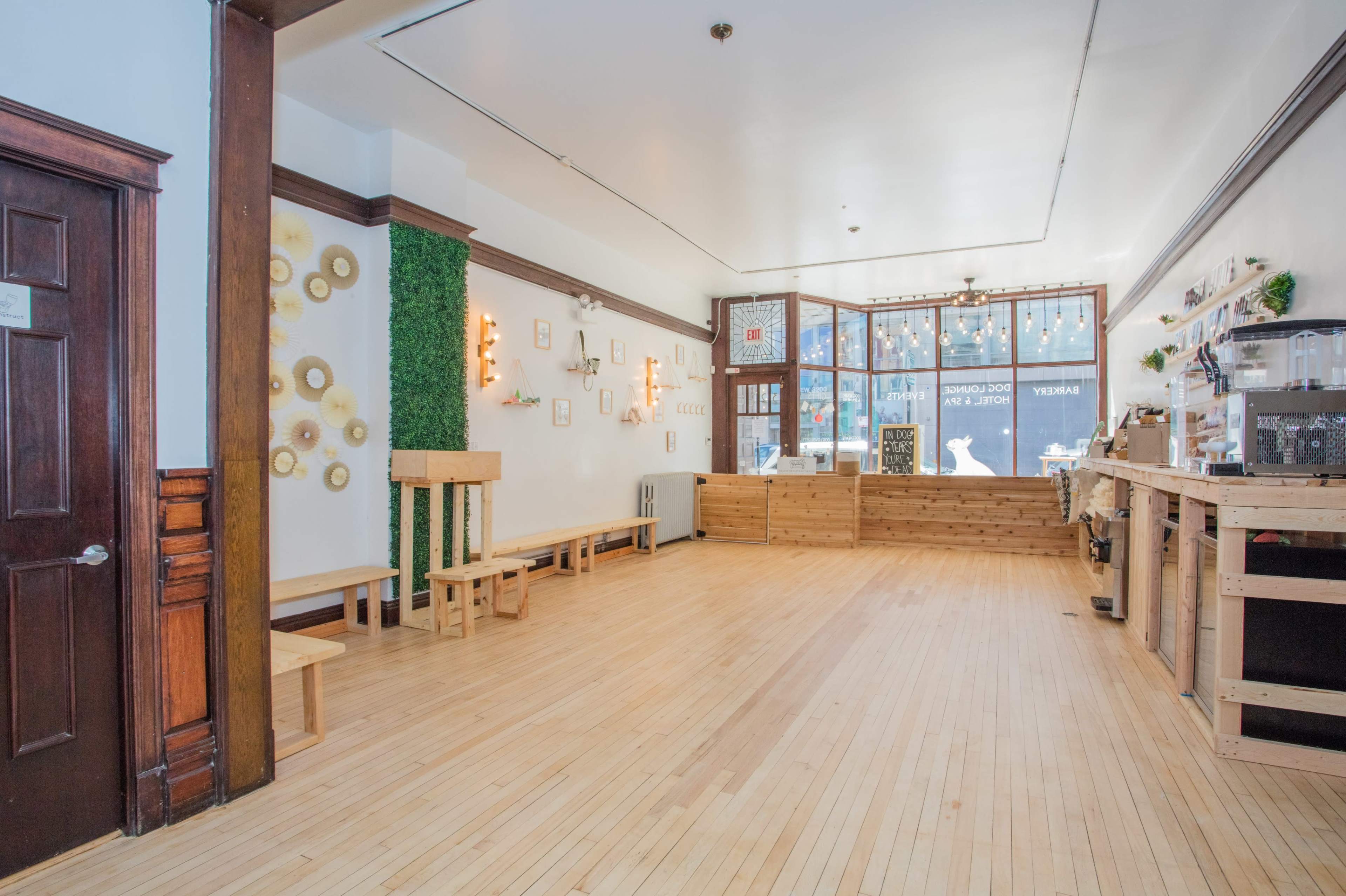 A spacious, empty room with wooden flooring, large front windows, and wooden benches along the walls.