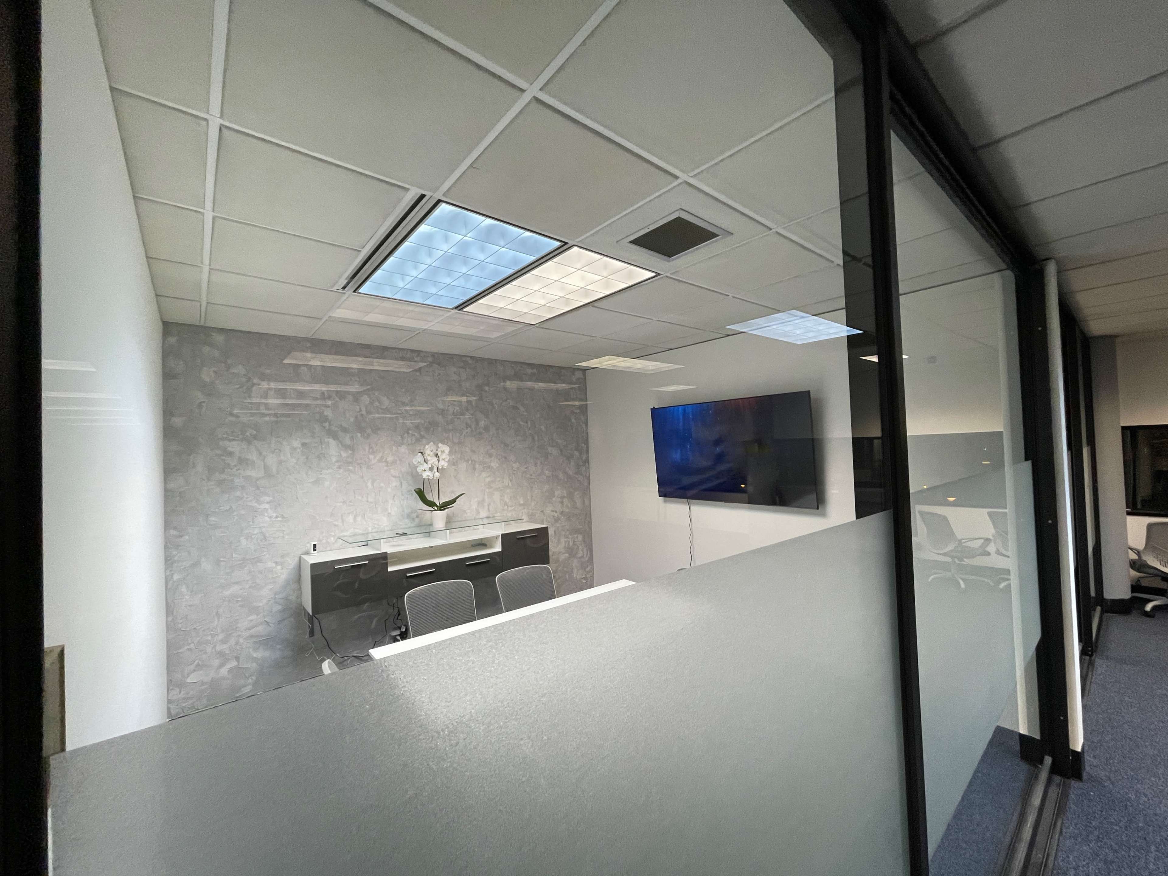A modern office conference room with a glass wall, featuring a table, chairs, and a mounted television.