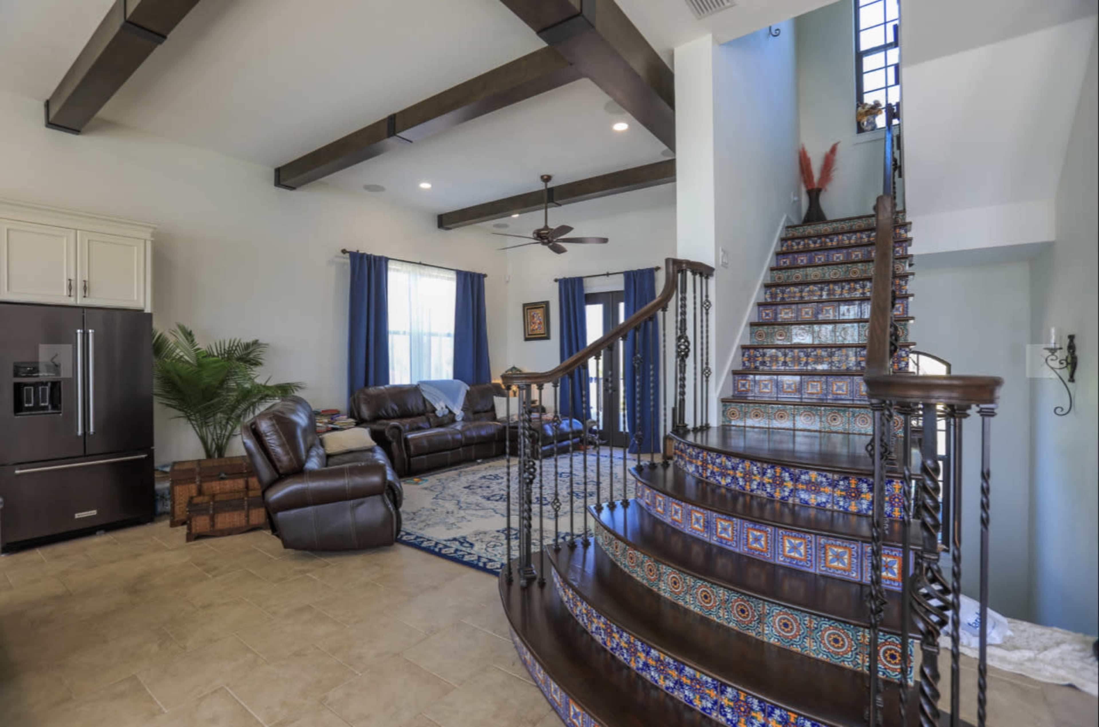 The image shows a spacious living room with a staircase featuring decorative tiles, a dark leather sofa set, and a large refrigerator in the kitchen area.