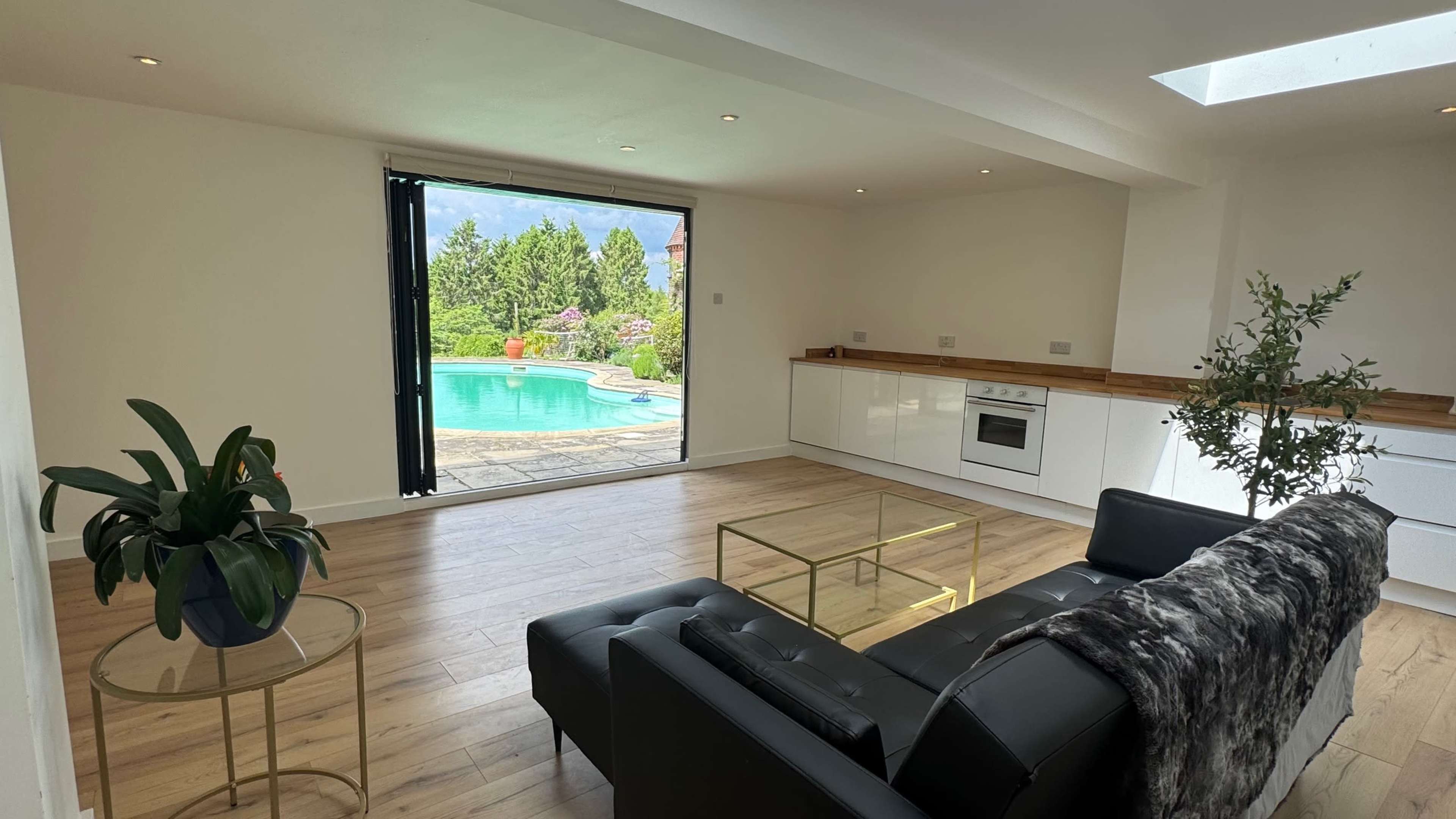 The image shows a modern living room with a black leather sofa, a glass coffee table, and a large window that opens to a patio with a swimming pool and landscaped garden.