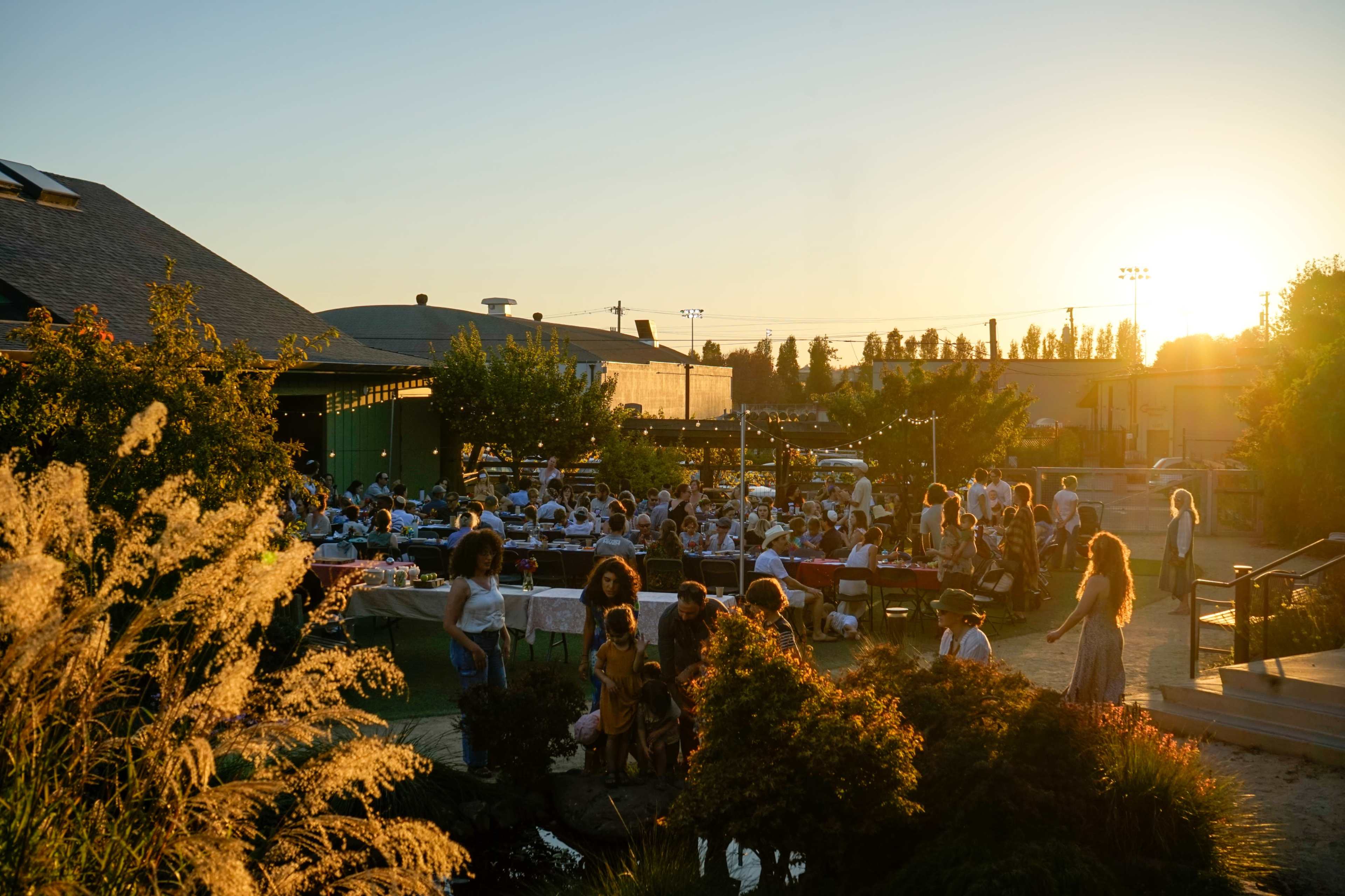 A large gathering takes place outdoors at sunset, with people seated at tables arranged around a garden.