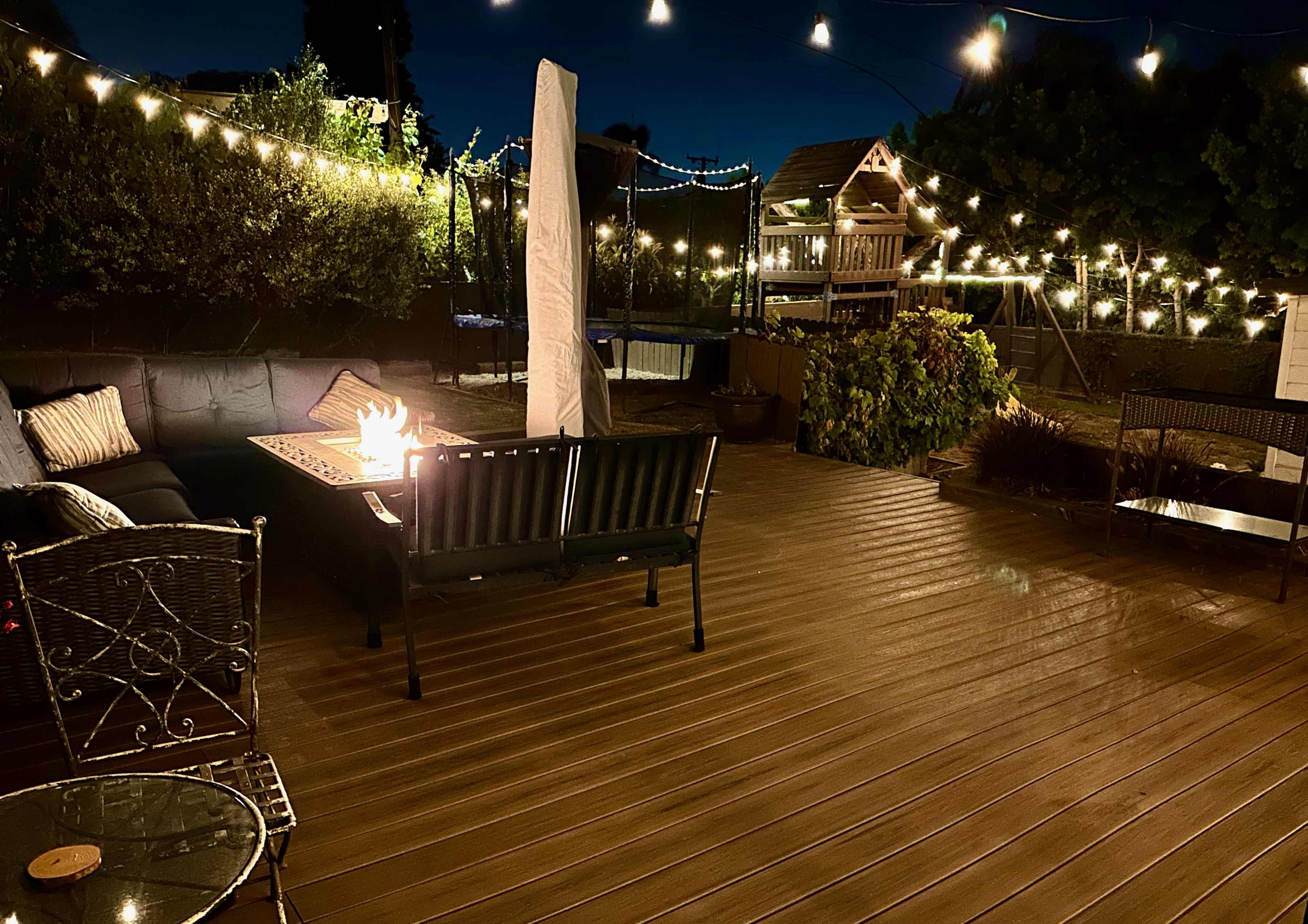 A wooden deck features a fire pit surrounded by seating, with string lights illuminating the area at night.