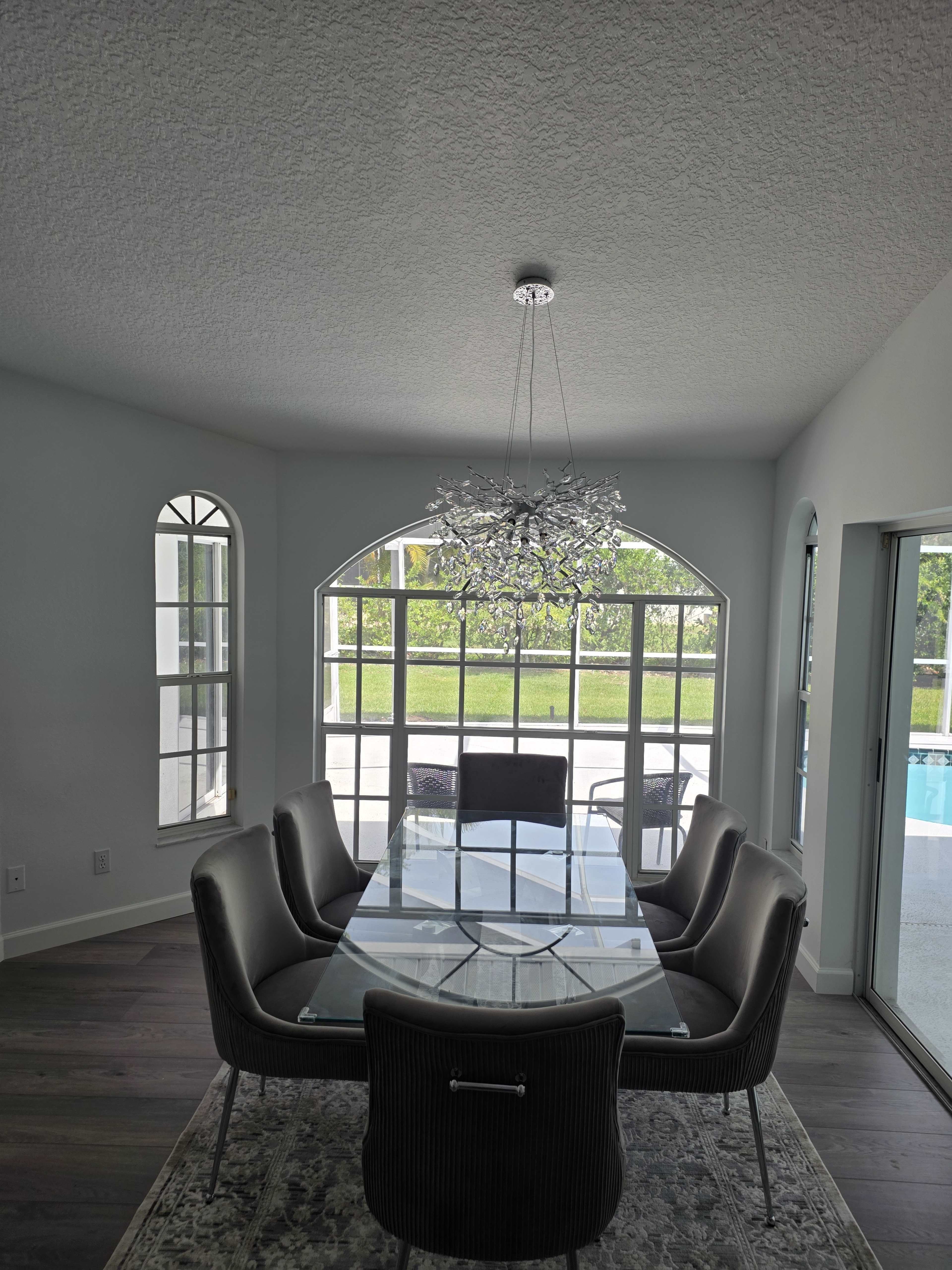 A modern dining room features a glass table surrounded by six upholstered chairs, with large windows providing a view of a pool area.